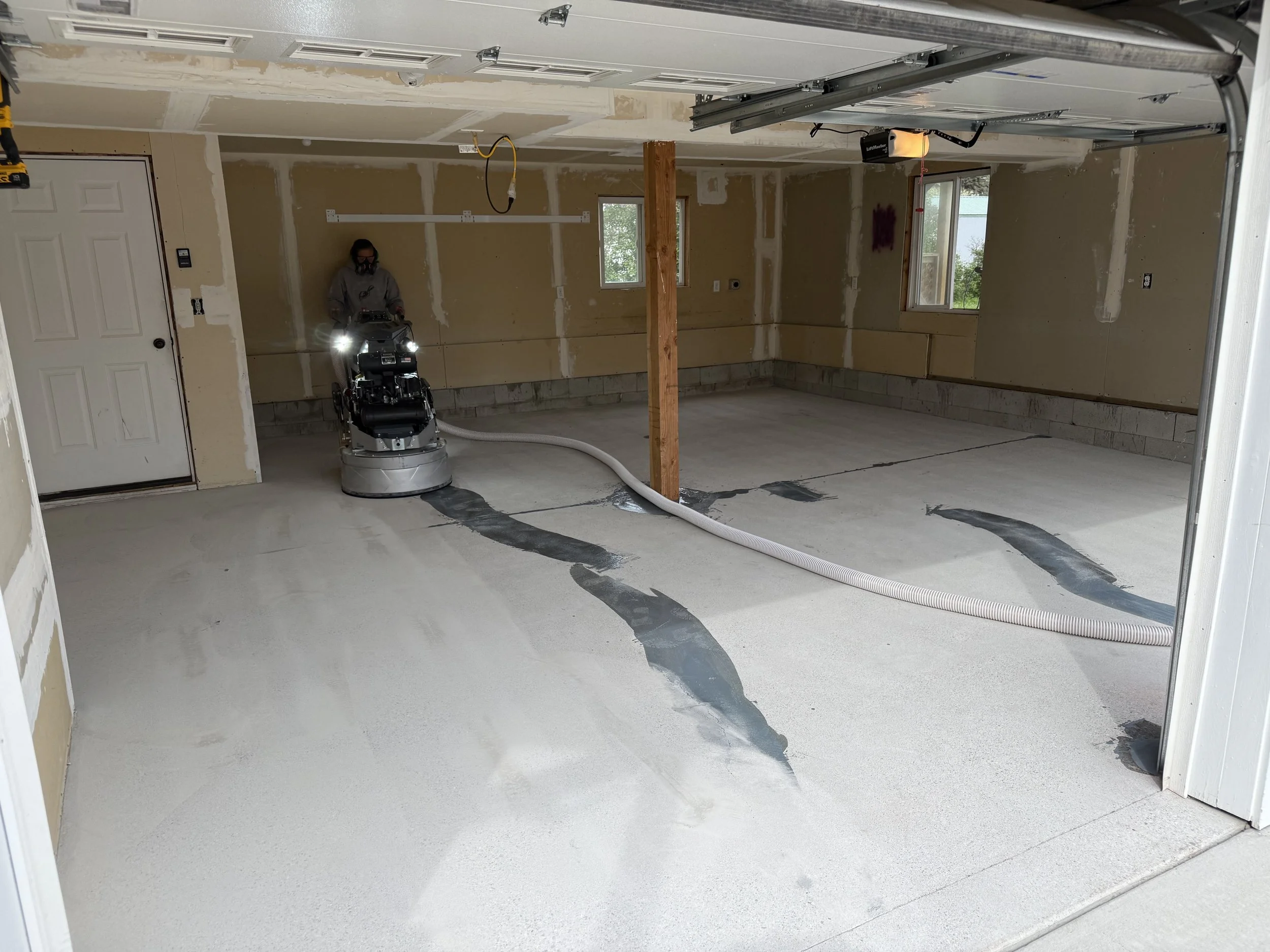 Garage under construction with a worker operating a floor grinding machine, concrete floor being leveled, exposed drywall, support beam, and a partially open garage door.