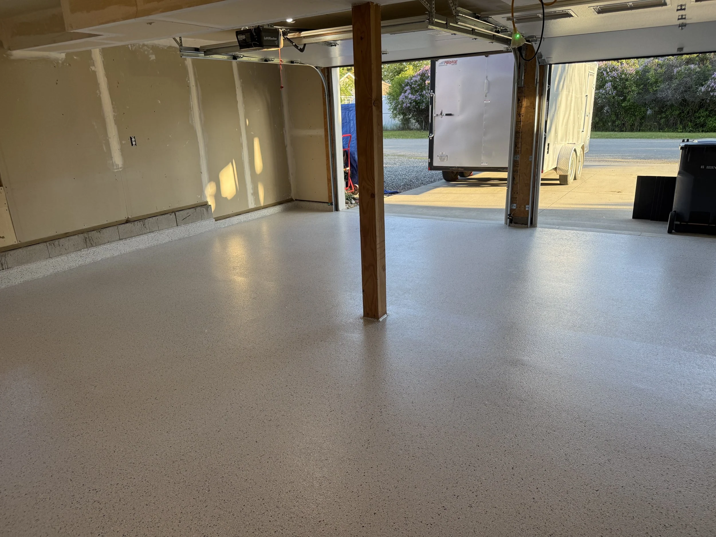 Empty garage with a newly finished epoxy floor, partially finished walls, and a view outside through the open garage door showing a trailer and greenery.