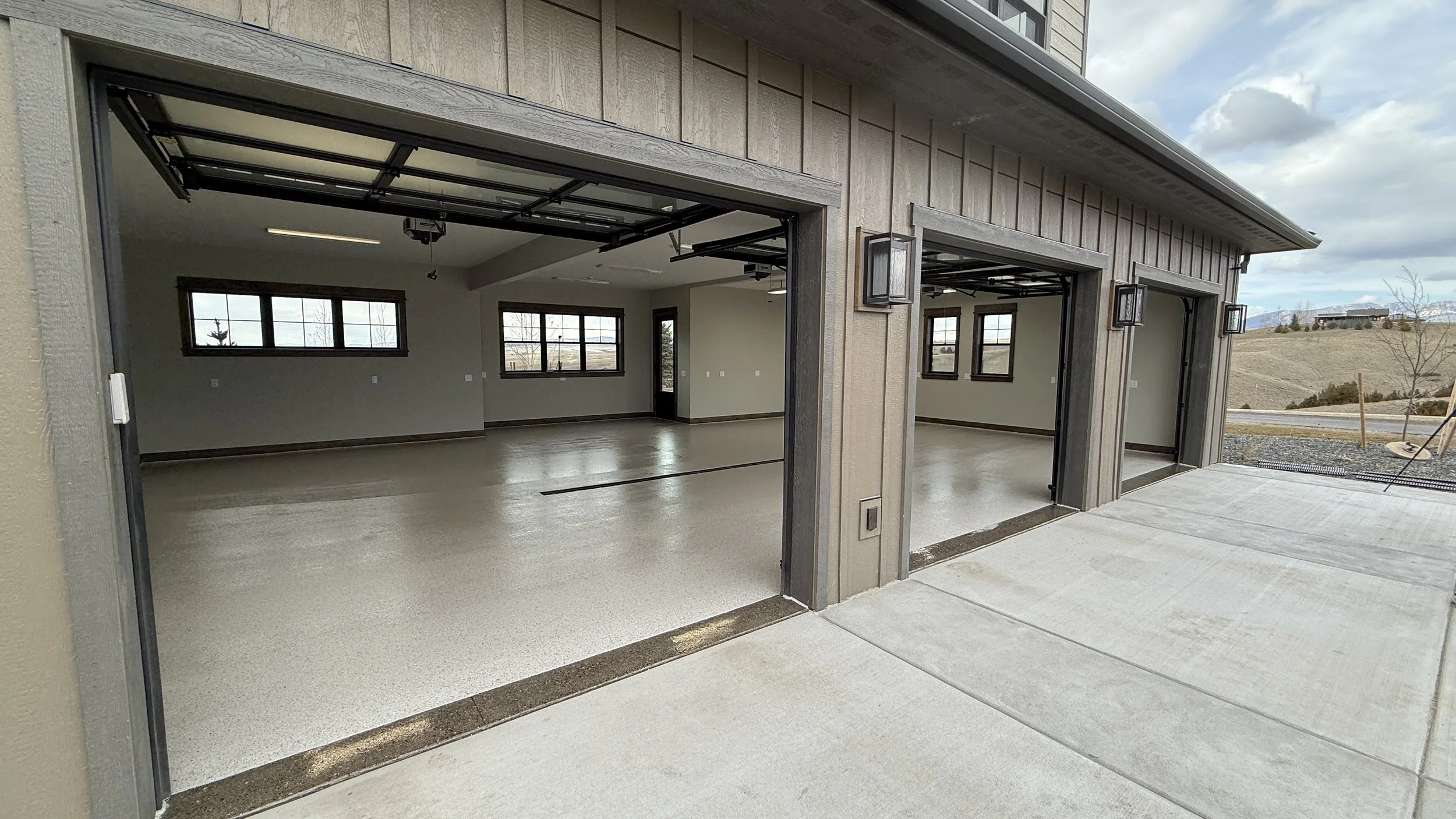 Empty two-car garage with closed white garage doors, gray walls, and overhead lighting. There are small windows above the garage doors and a small door with a window on the right side.