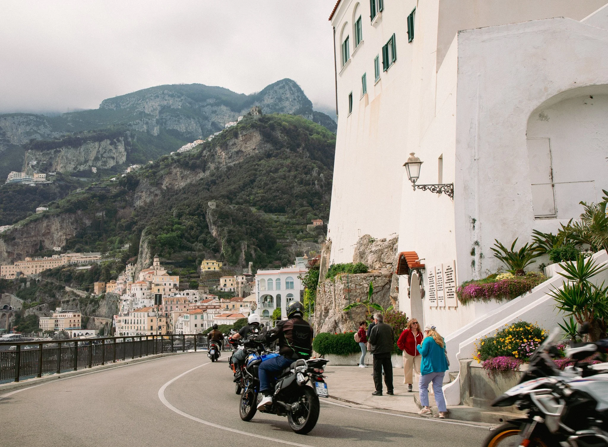 Tourists and motorcyclists gather near a coastal walkway with a white building and mountain village in the background.
