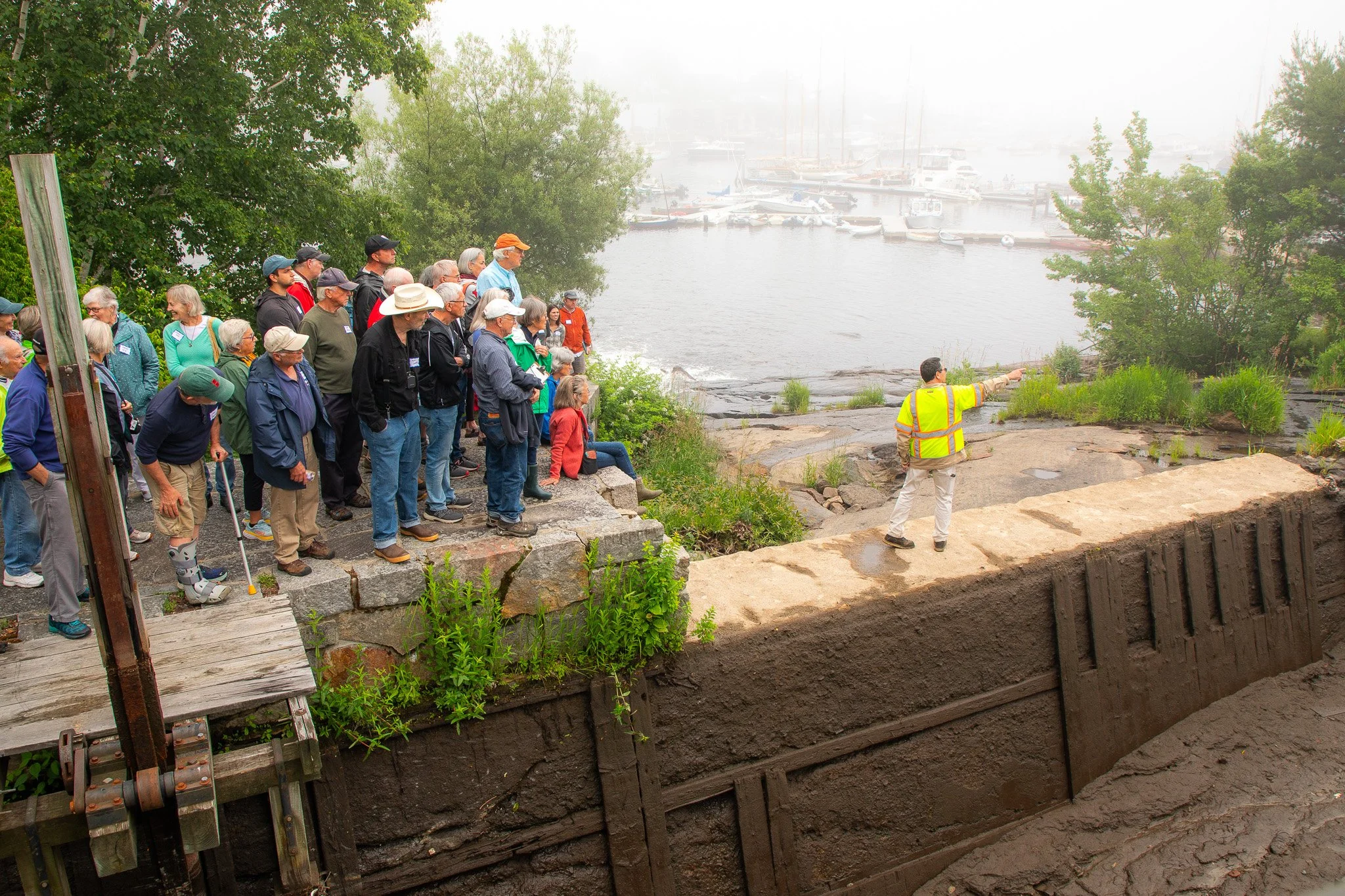 A group of people observing a construction site near a waterfront with boats, guided by a person in a high-visibility vest.