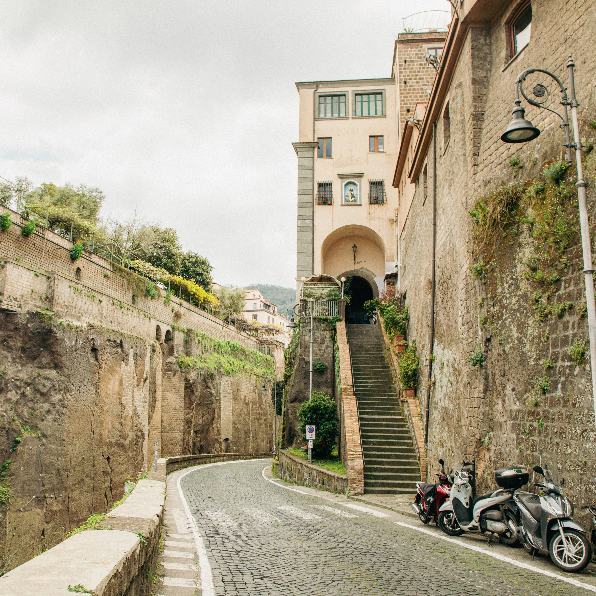 A narrow cobblestone street with parked scooters on the right side, leading past old stone and brick buildings with steps and greenery nearby, overcast sky.