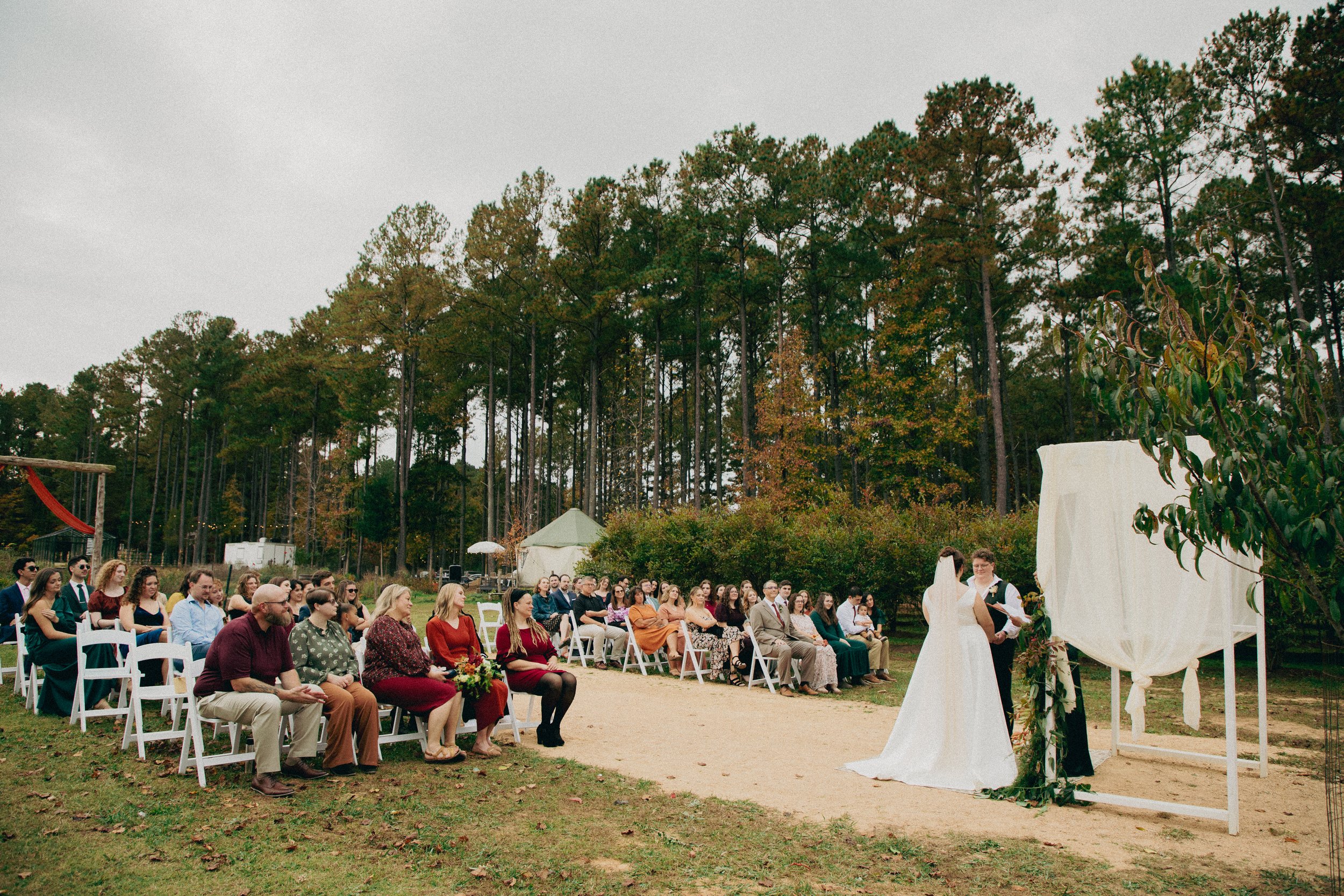 An outdoor wedding ceremony with seated guests, a bride and groom exchanging vows, and a forest background under an overcast sky.