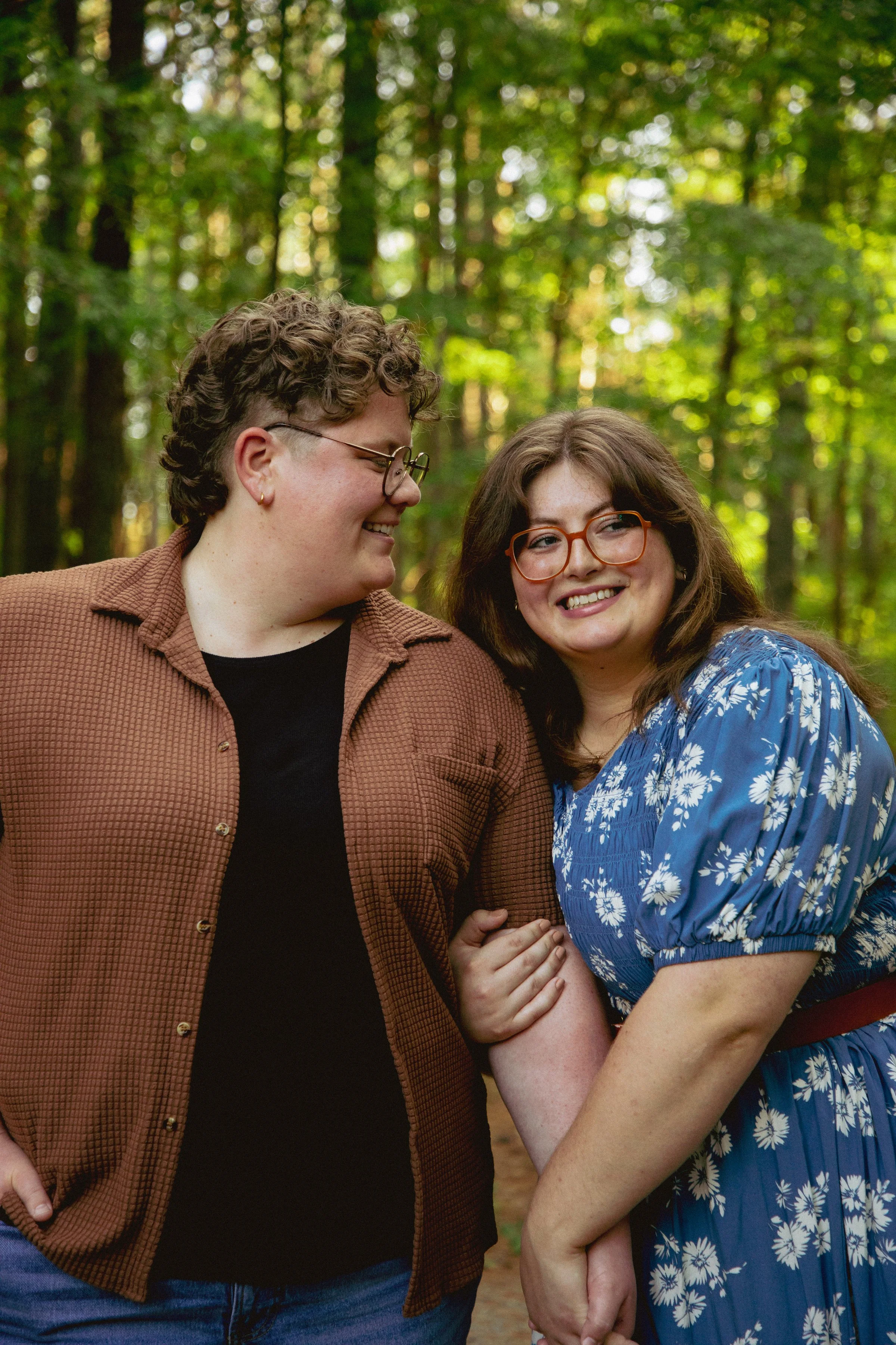 Two women standing close together in a forest, holding hands, smiling, and looking at each other. One woman has short curly hair, glasses, and is wearing a brown shirt. The other woman has long hair, glasses, and is wearing a blue dress with white fl