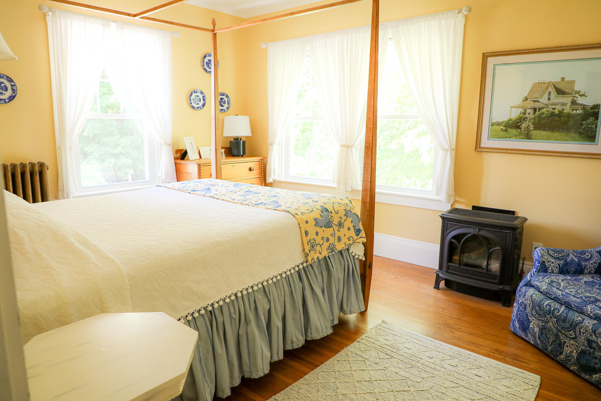 A cozy bedroom with yellow walls, a large bed with a pale yellow quilt, a wooden bedside table with a lamp, and a picture of a house on the wall. Natural light floods in through multiple windows with white curtains. There is a blue patterned armchair