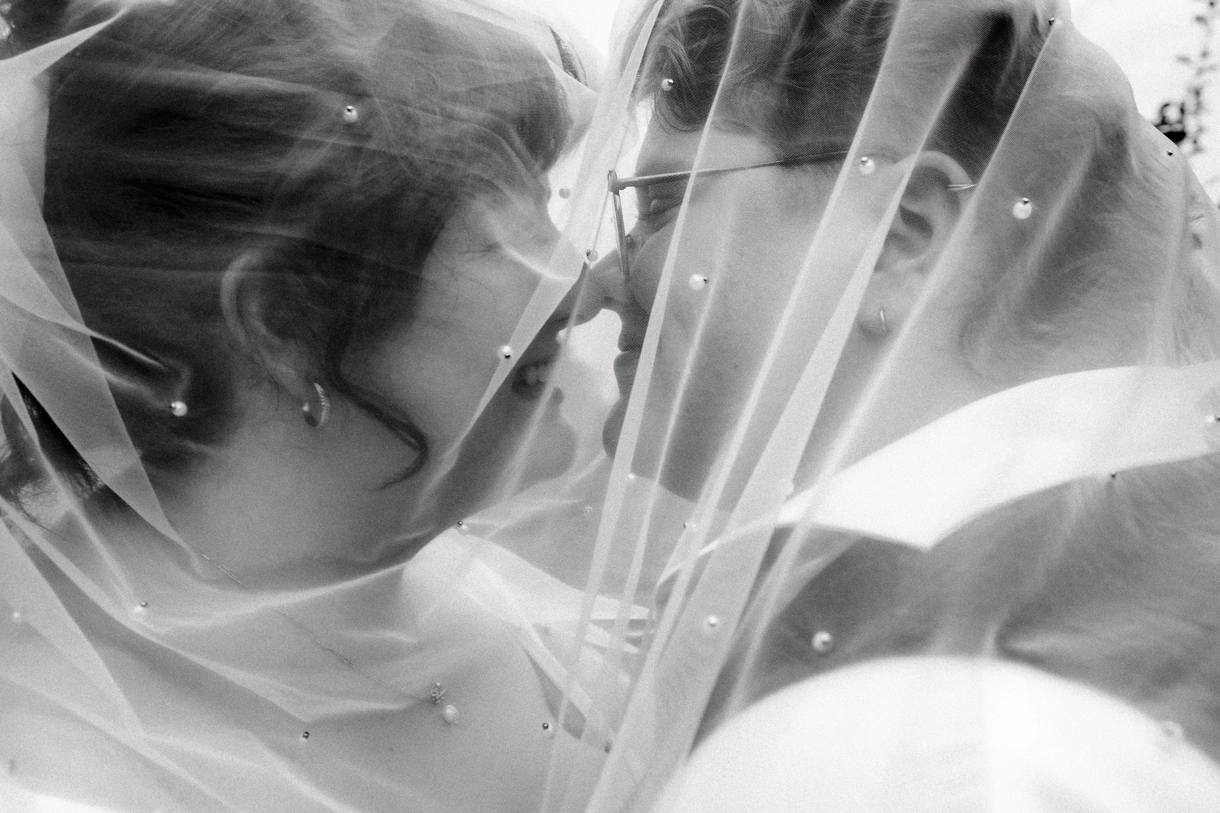 A couple dressed in protective clothing and old-fashioned glasses are facing each other closely, behind a transparent barrier with small metal fasteners, in black and white.