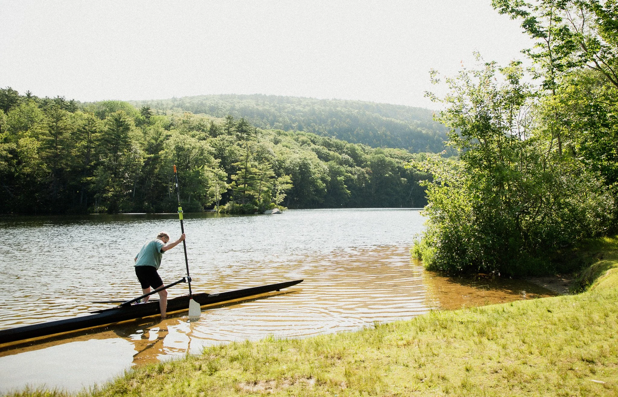A person preparing to paddleboard on a calm lake surrounded by lush green trees and rolling hills.