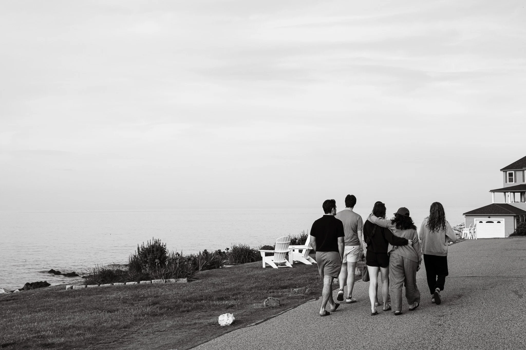 A group of six people walking along a seaside path with houses and chairs nearby, overlooking a large body of water with a cloudy sky overhead.