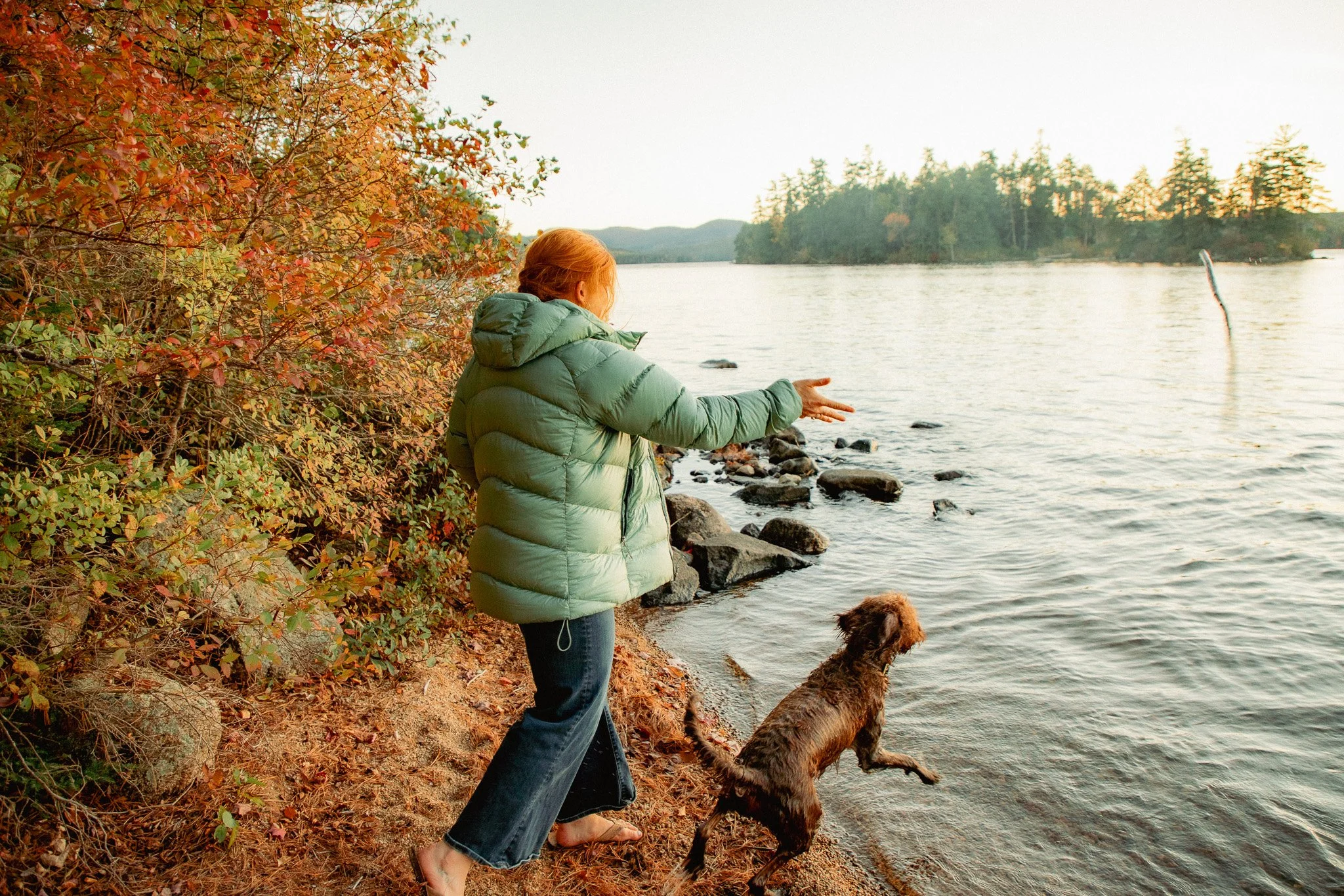 A woman with red hair in a green puffy jacket and jeans walking her dog along a rocky shoreline by a lake during autumn, with trees displaying fall colors and hills in the background.