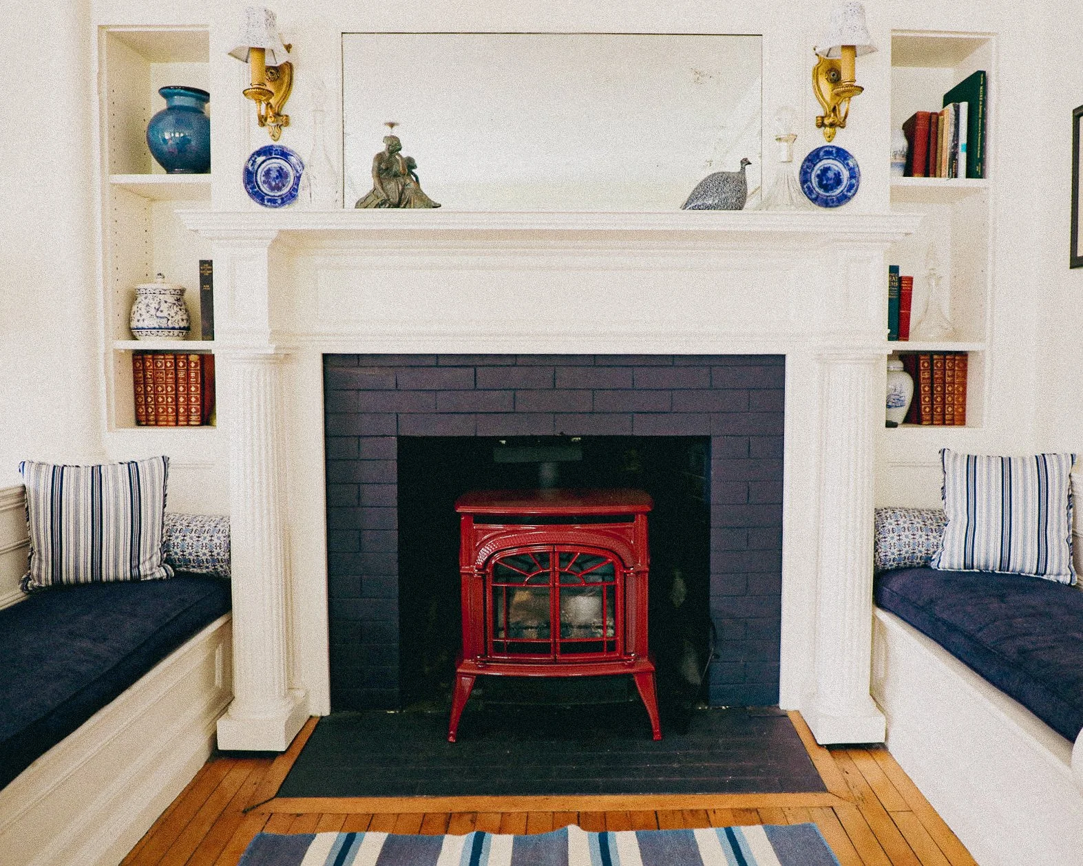 Living room fireplace with built-in bookshelves on each side, decorated with vases, books, and statues, with a red wood stove inside the black brick fireplace, flanked by built-in seating with striped and patterned pillows, and a striped rug on woode