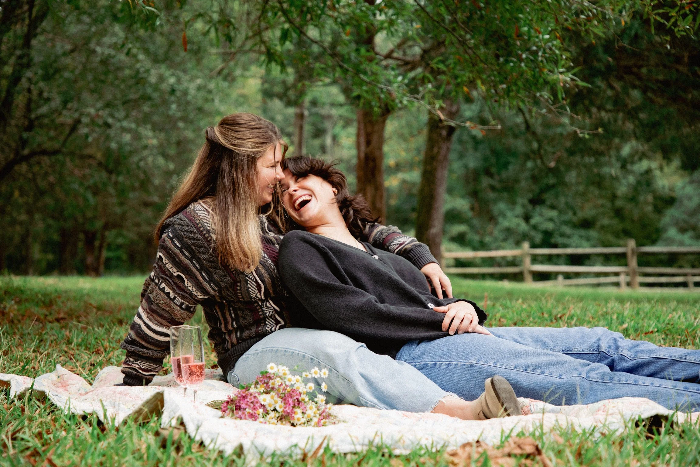 Two women enjoying a picnic on a blanket in a park with green trees in the background. One woman is sitting with her head back, smiling, while the other woman leans in close, also smiling.