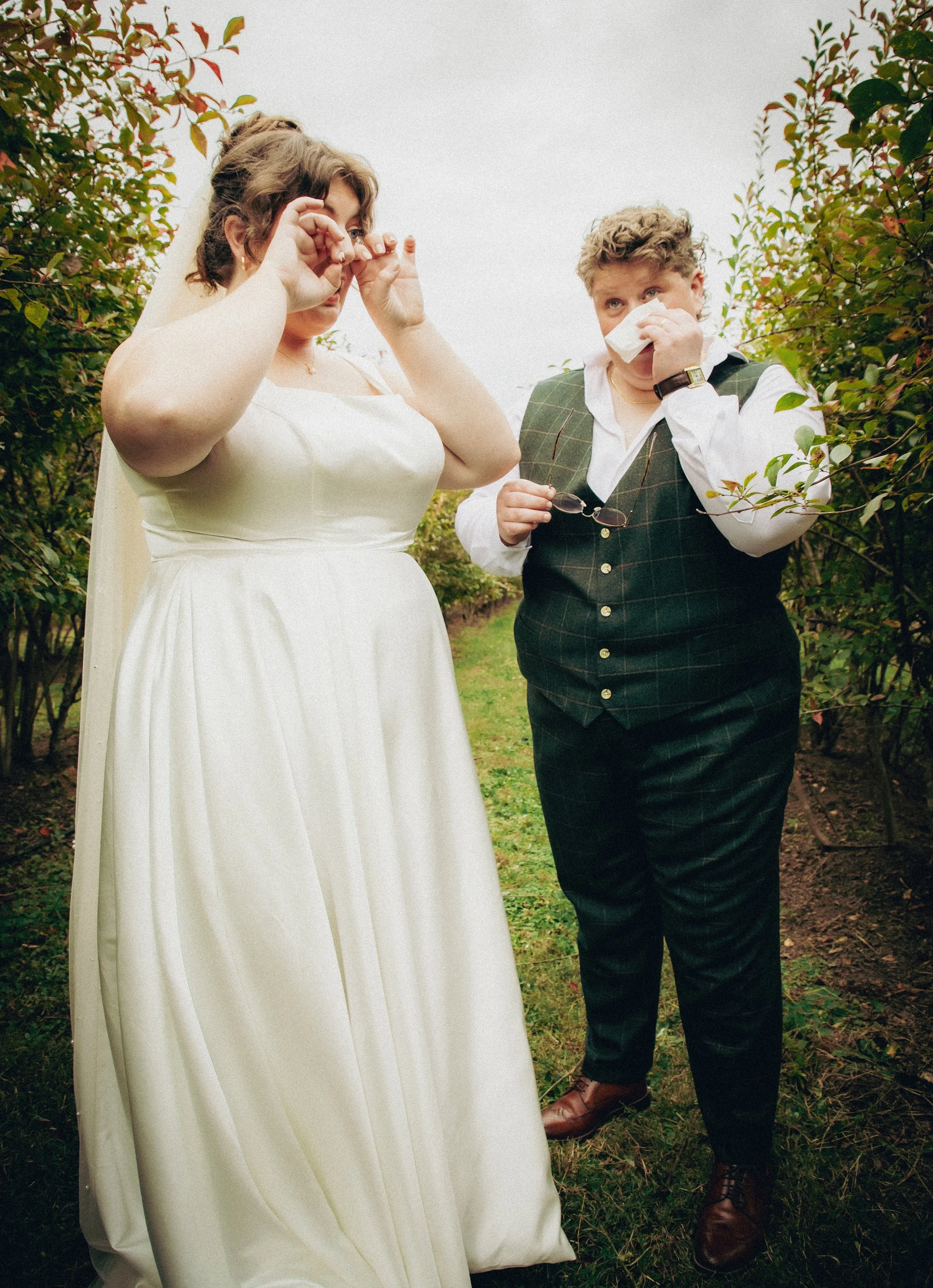A bride in a white wedding dress and a groom in a dark plaid vest and trousers stand outdoors among green bushes. The groom is with a tissue, and the bride is adjusting her glasses or ear.