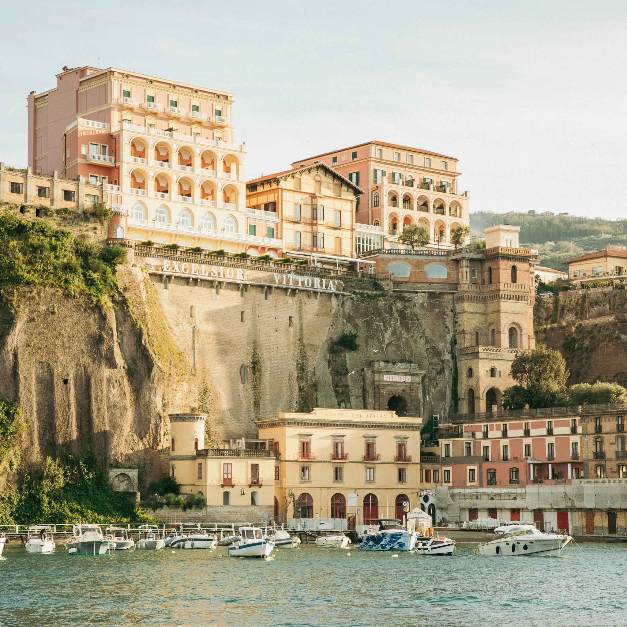 Marina with several white boats docked in the water in front of pastel-colored buildings on a hillside.