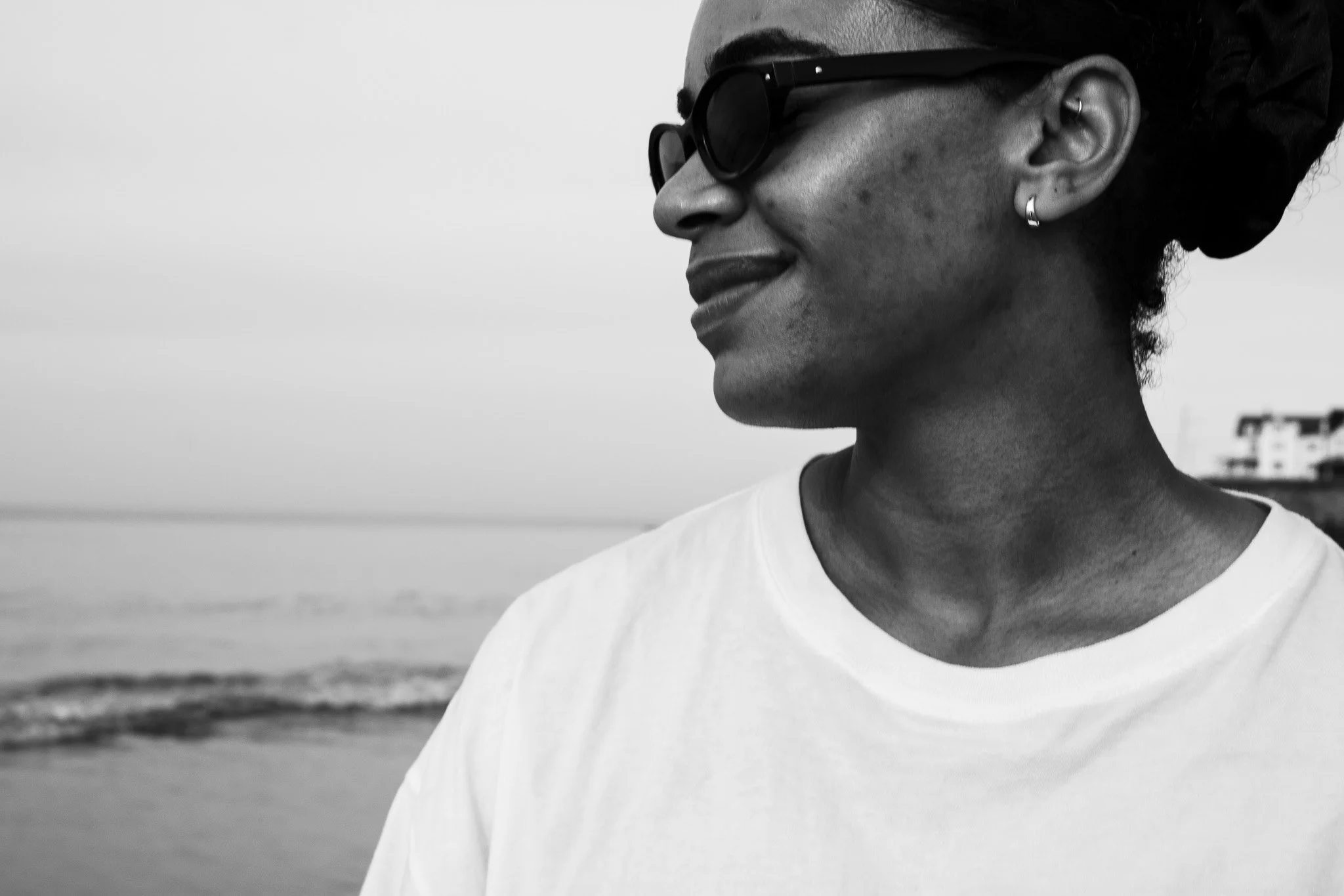 A woman with dark hair styled in a bun, wearing dark sunglasses and small hoop earrings, smiling and looking to the side, on a beach with buildings in the background, in black and white.
