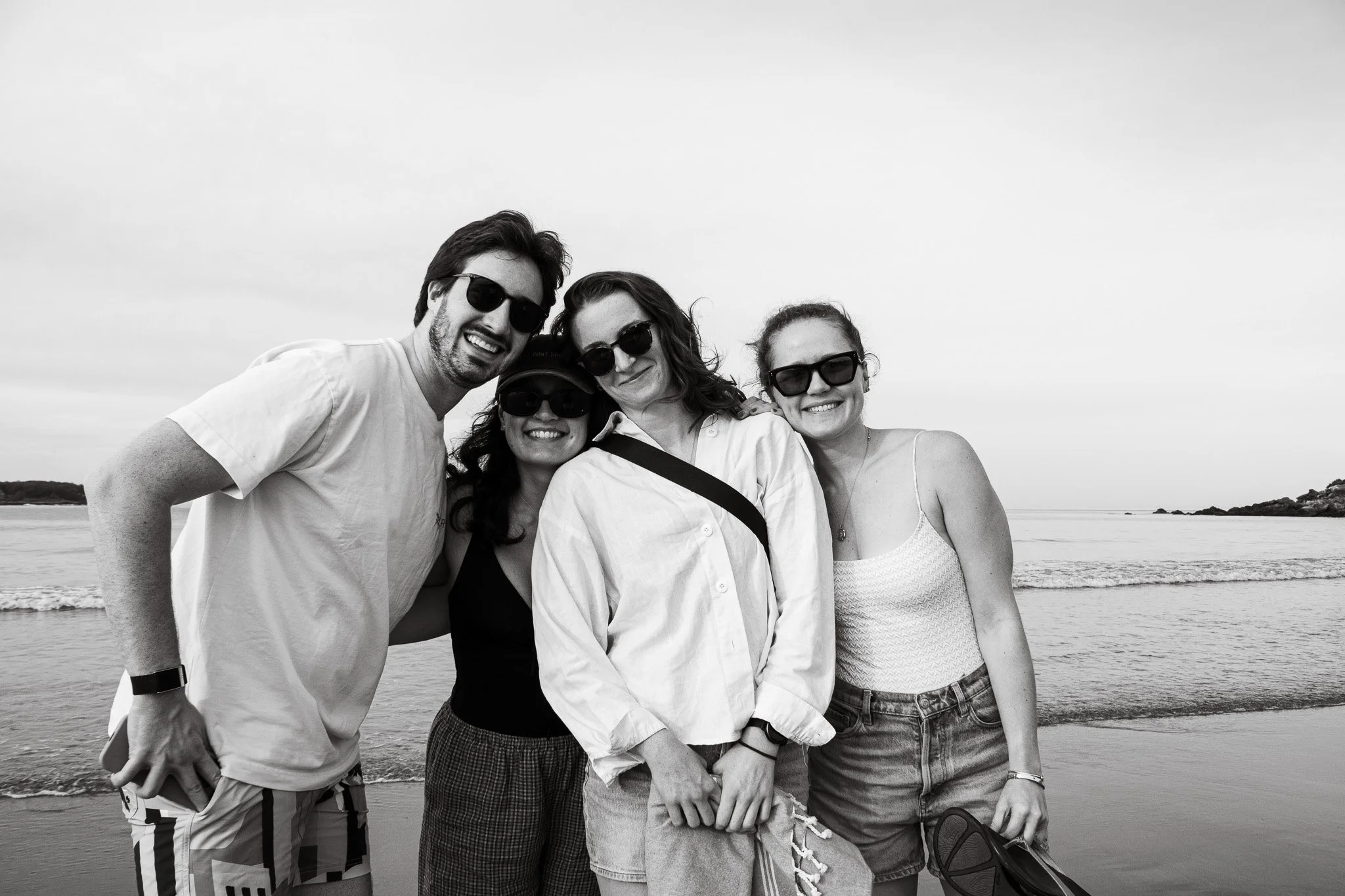 Four friends standing together on the beach, smiling and wearing sunglasses, with the ocean and shoreline in the background.