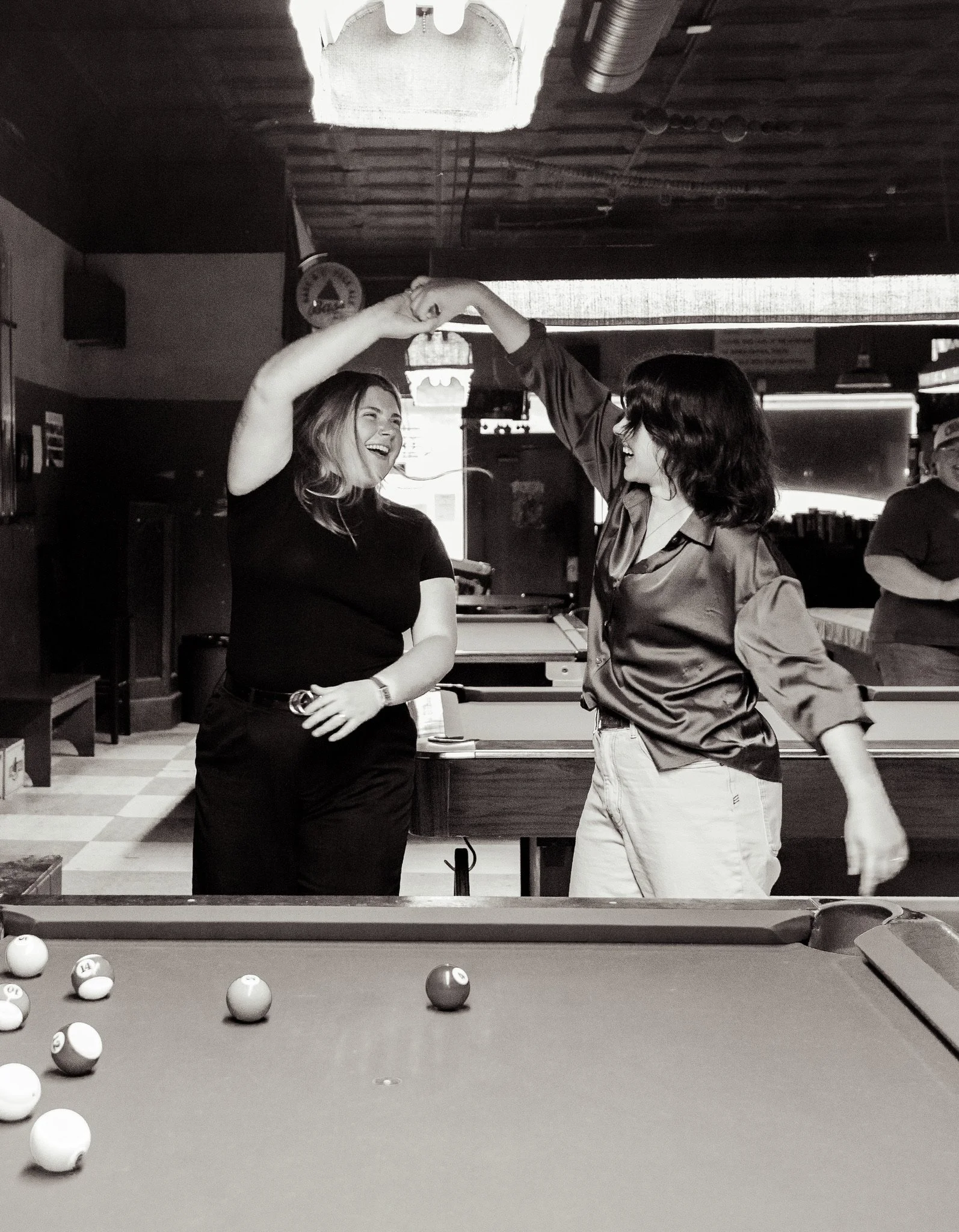 Two women are smiling and holding hands in a dance pose in front of a pool table, with billiard balls scattered on the table. The setting appears to be a bar or pub.
