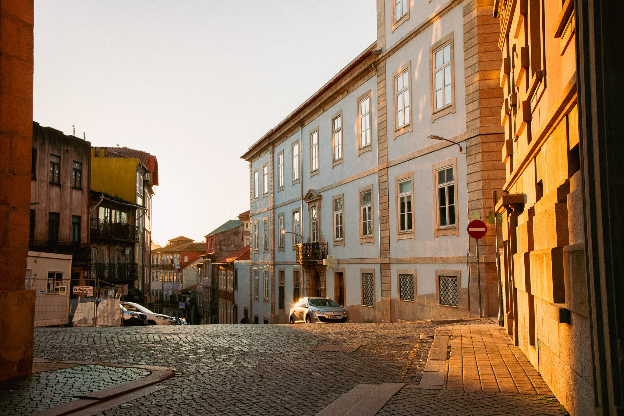 A quiet city street at sunset with cobblestone pavement, colorful buildings, and parked cars.