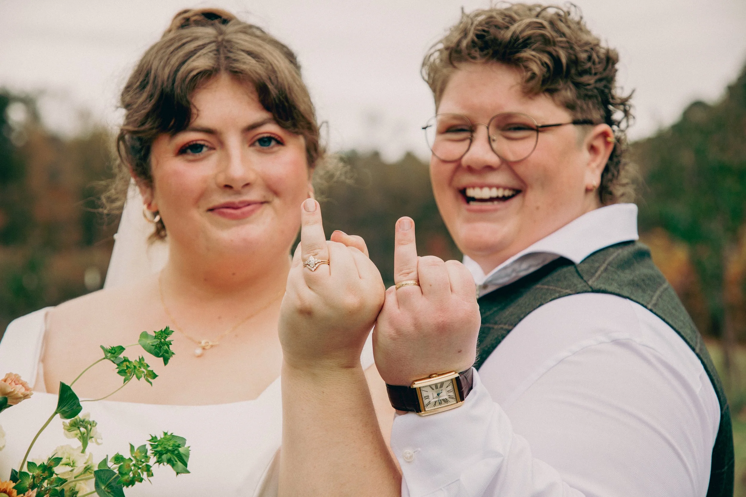 A man and woman, both smiling and showing their middle fingers, are close together outdoors during the daytime. The woman is wearing a wedding dress and jewelry, while the man has curly hair, wears glasses, a shirt, and a vest. The background is blur
