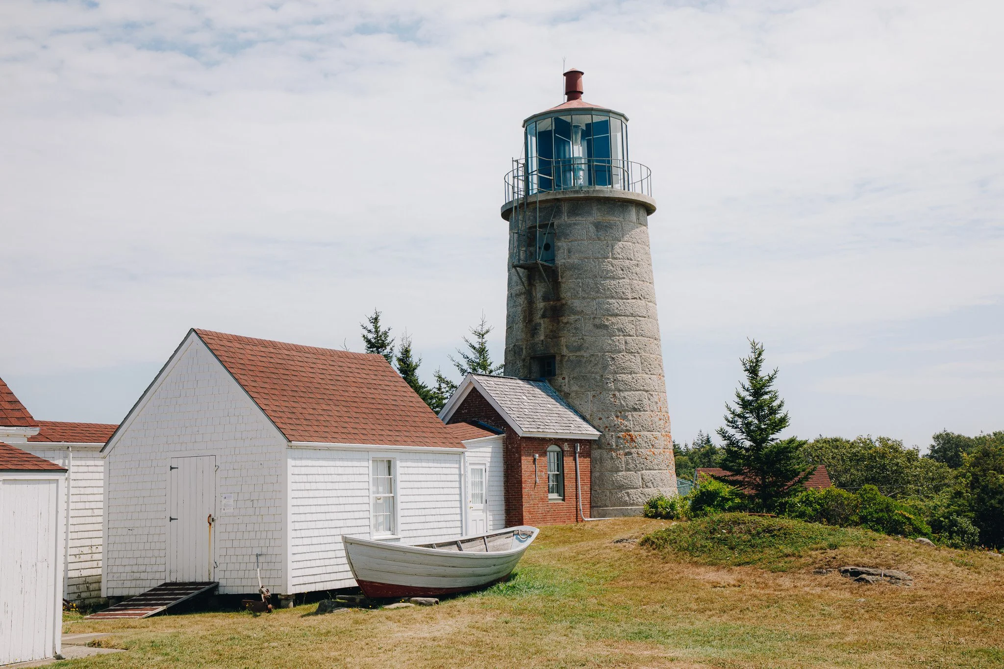 A lighthouse with a stone base and a blue glass enclosure at the top, surrounded by houses with red and white shingled roofs, a small boat on the grass, and trees in the background under a partly cloudy sky.