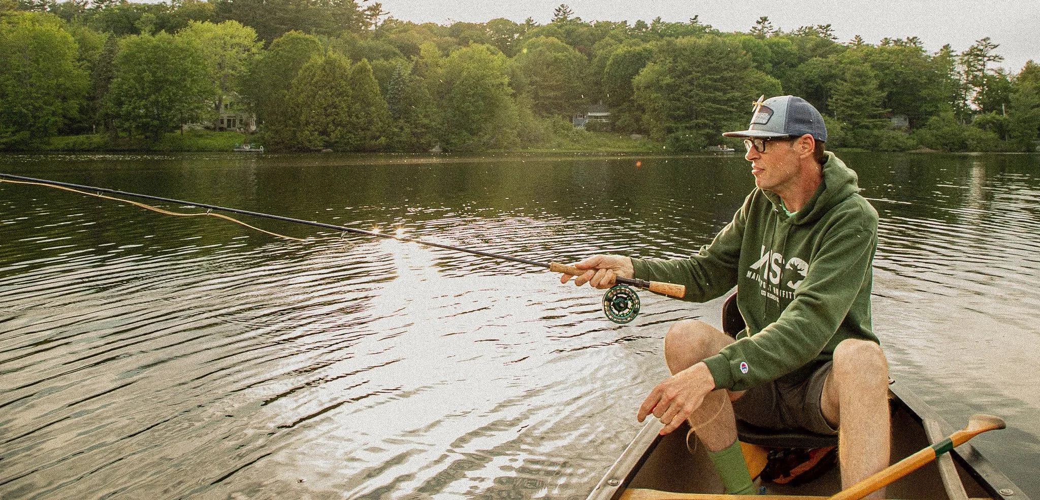 A man in a dark baseball cap, sunglasses, and green hoodie sitting in a boat on a lake, fishing with a fishing rod. The lake is surrounded by trees and houses in the background, and the water is rippling as the man reels in his catch.