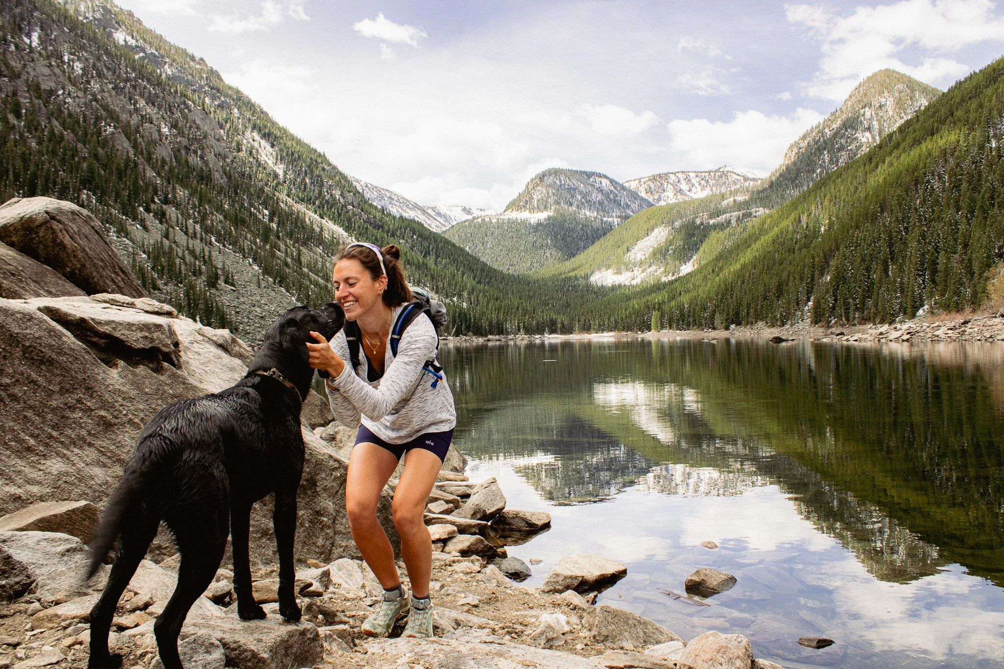 A woman smiling and playing with a black dog by a lake in a mountainous forest area on a partly cloudy day.