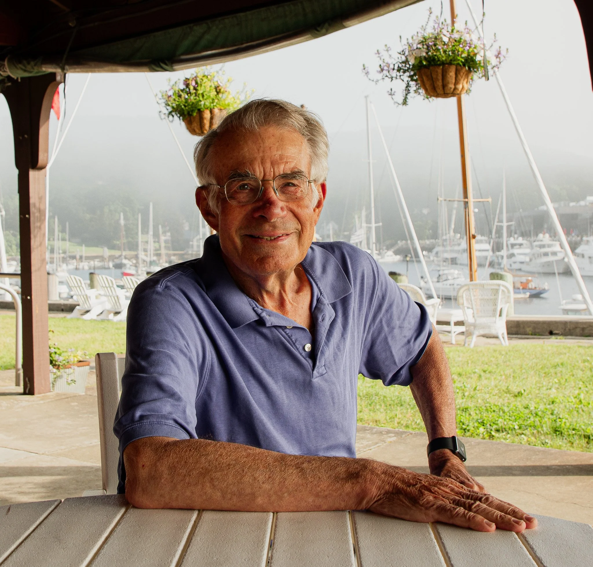 A man with glasses is doing a yoga pose on a white wooden table outdoors with boats and hills in the background.