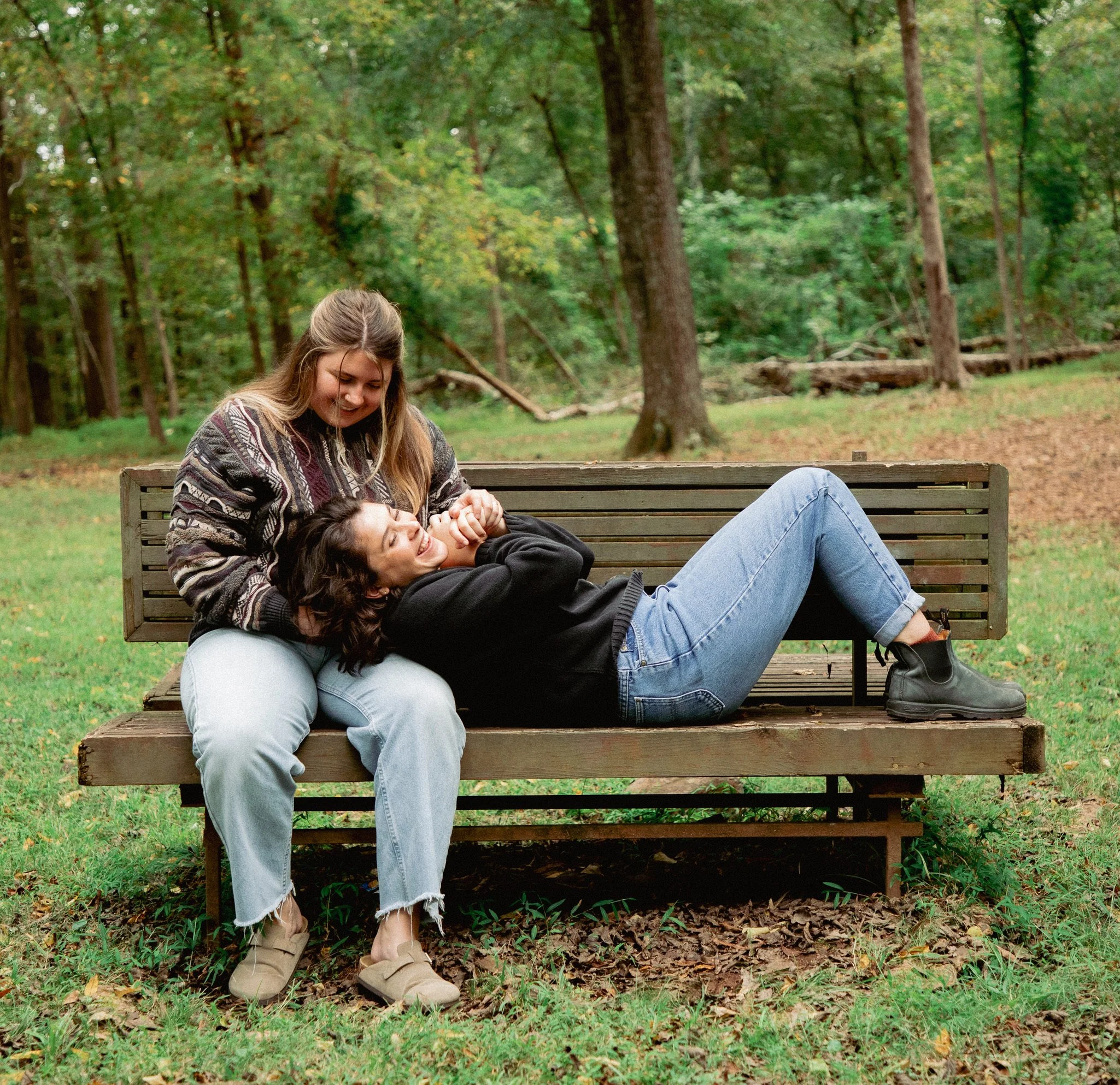 Two young women smiling and lying together on a park bench surrounded by trees and fallen leaves in a forest setting.