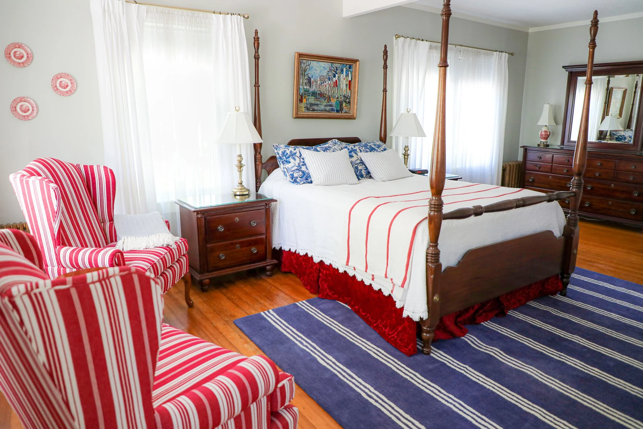 A bedroom with four-poster bed, striped armchair, blue and white rug, framed artwork, and furniture including nightstands and dresser.
