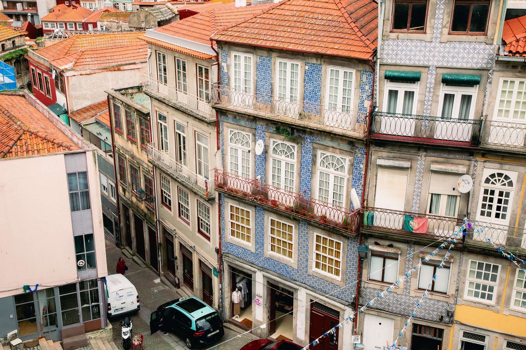 A view of colorful old European-style buildings with tiled roofs, balconies, and narrow streets, featuring vibrant facades, laundry hanging, and parked cars.