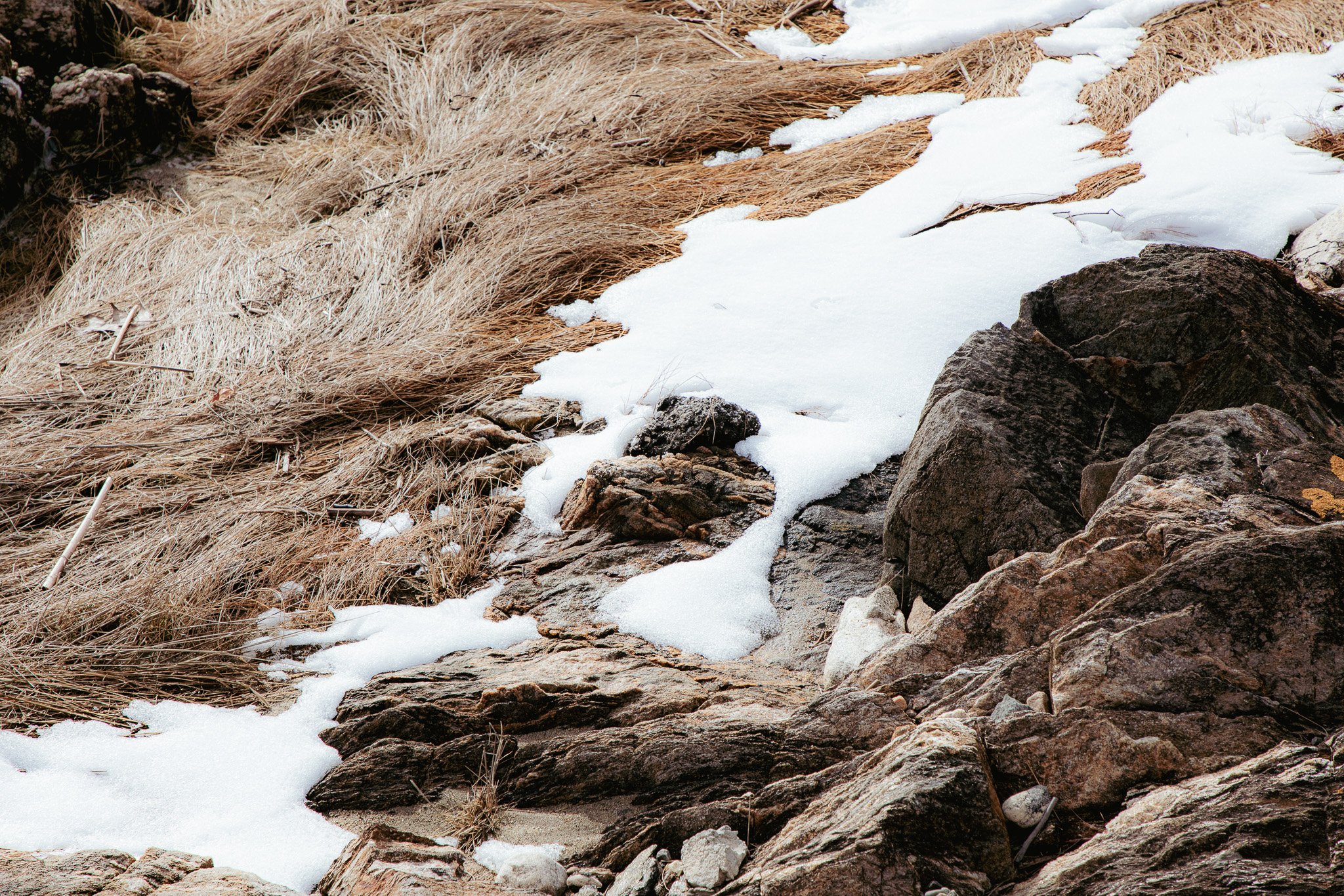 Frozen stream flowing through rocks and dry brown grass with patches of snow.