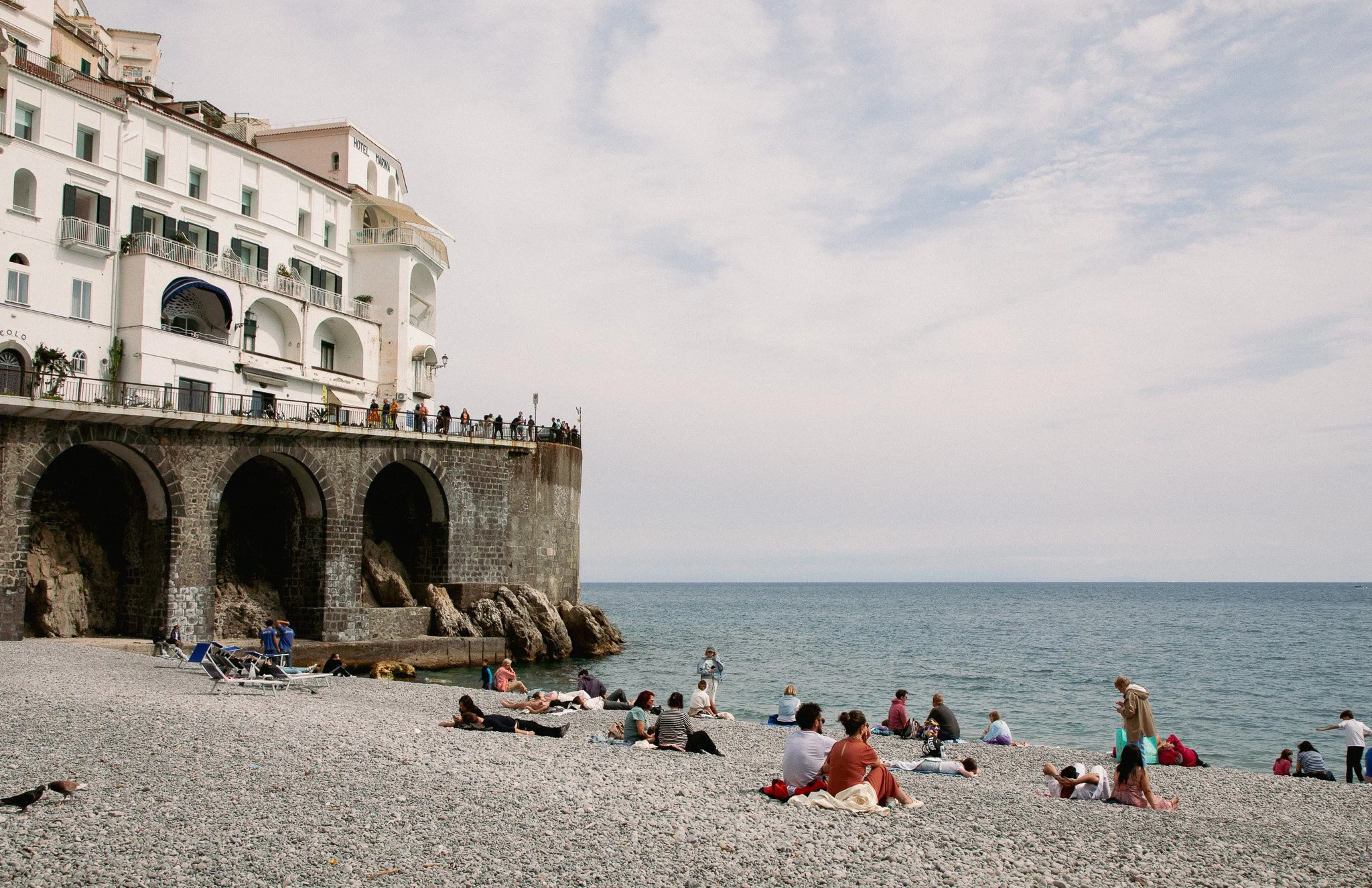People relaxing on a pebble beach by the ocean with a large white building on stilts in the background and a partly cloudy sky overhead.