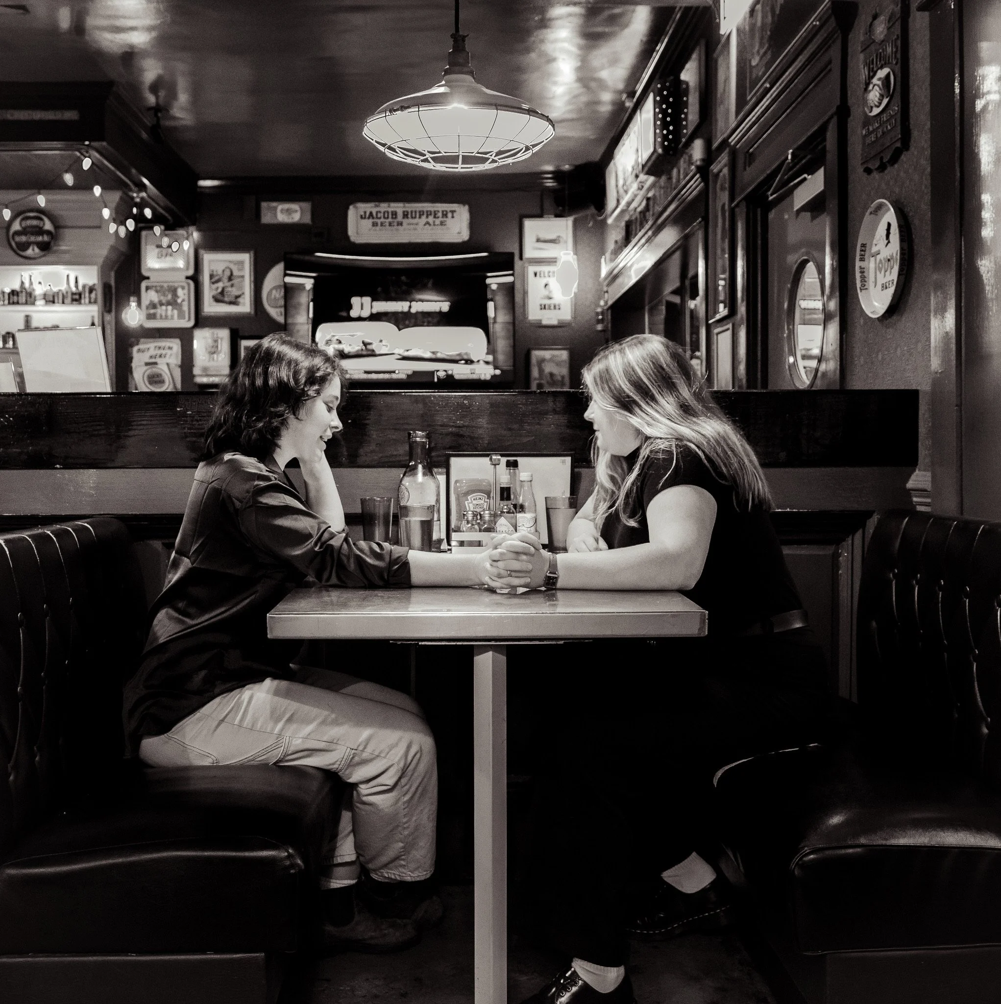 Two women sitting across from each other at a restaurant table, arm wrestling and smiling. The restaurant has vintage decor, framed pictures, and signs on the walls. Overhead light fixture is visible.
