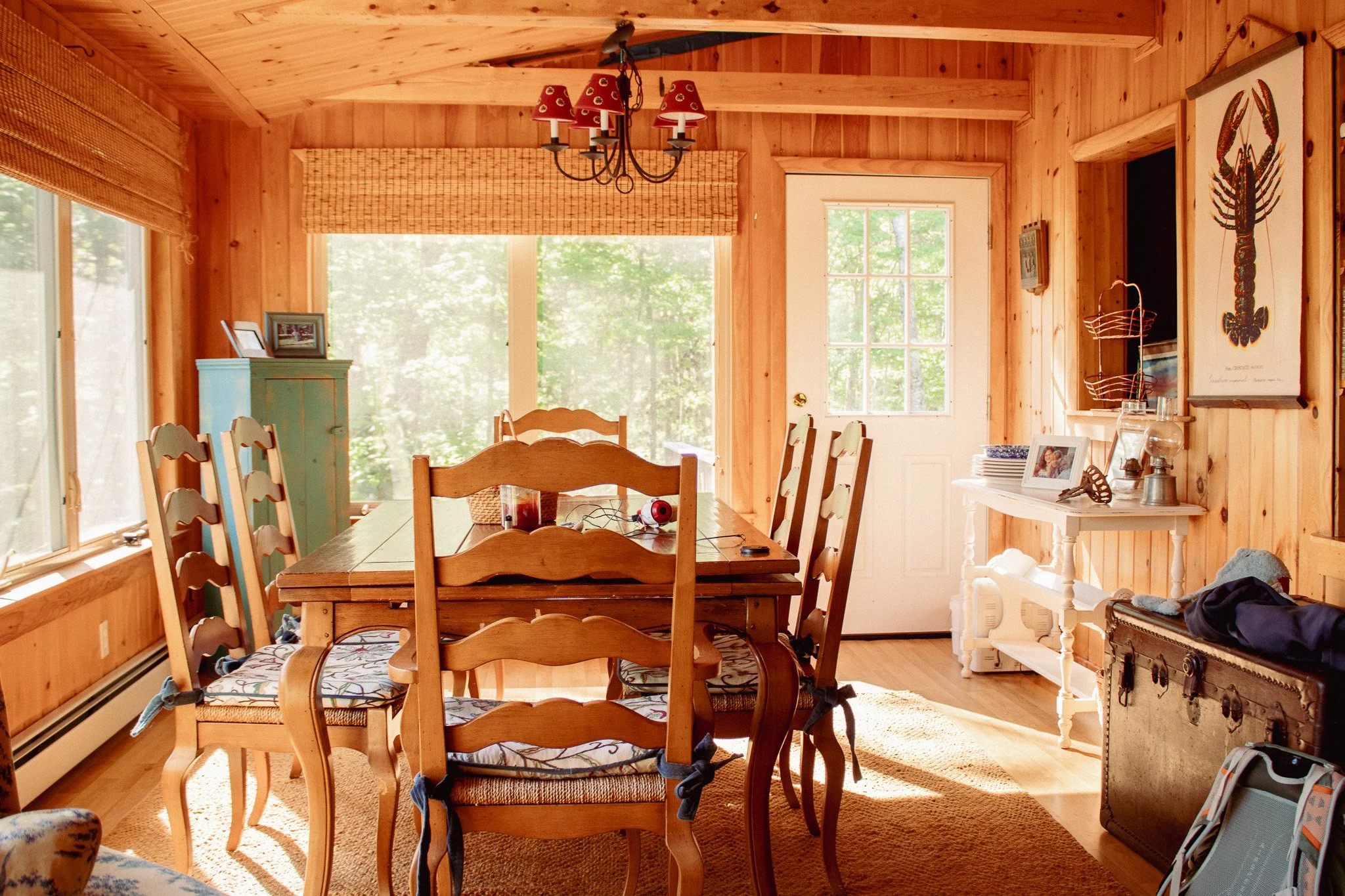 Wood-paneled dining room with large windows, a dining table with chairs, a chandelier with red lampshades, and various framed pictures and decorative items on shelves.