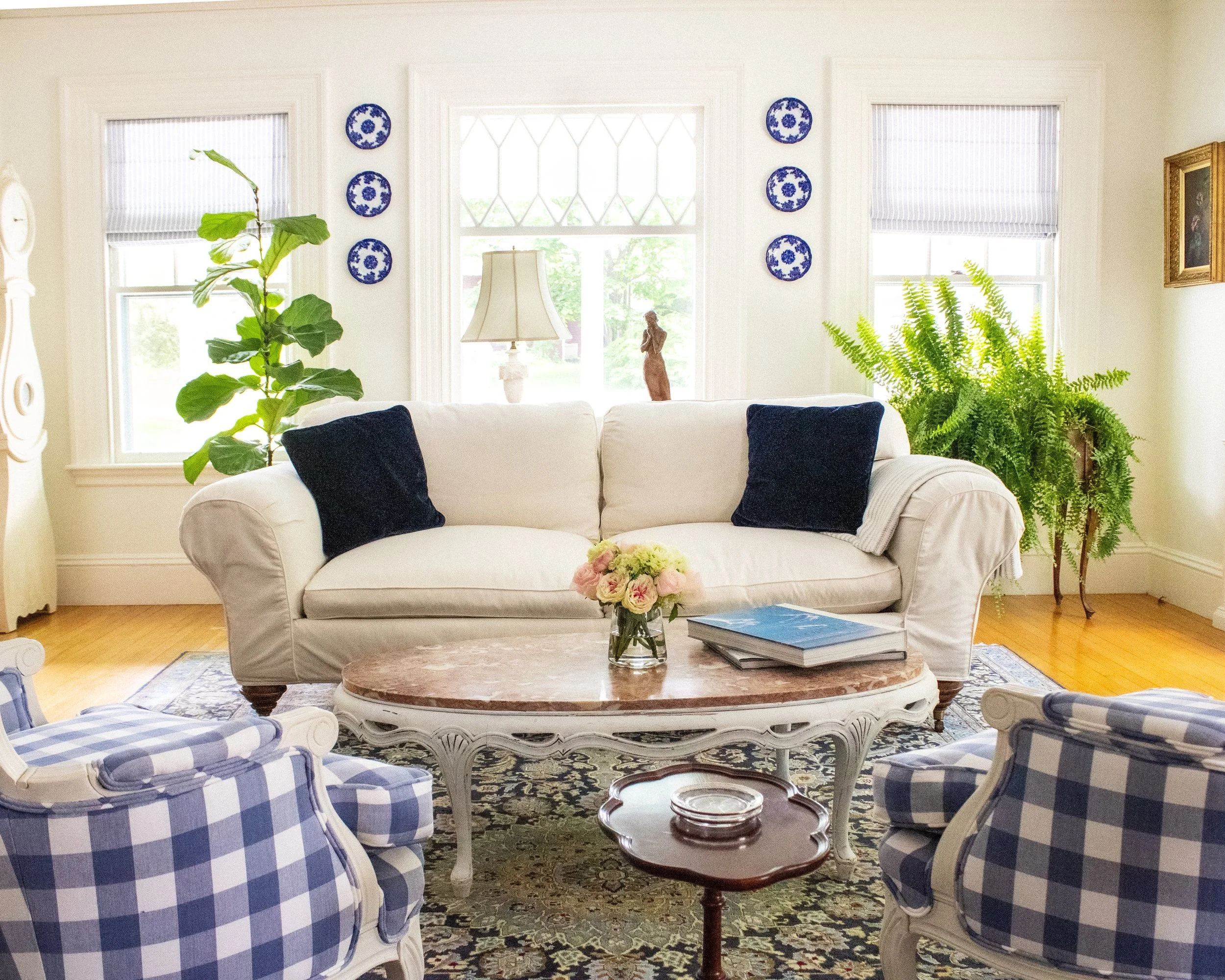 A living room with a white sofa with dark blue pillows, circular marble coffee table, blue and white plaid armchairs, greenery including a potted plant and fern, decorative plates on the wall, windows with striped blinds, a table lamp, a small sculpt
