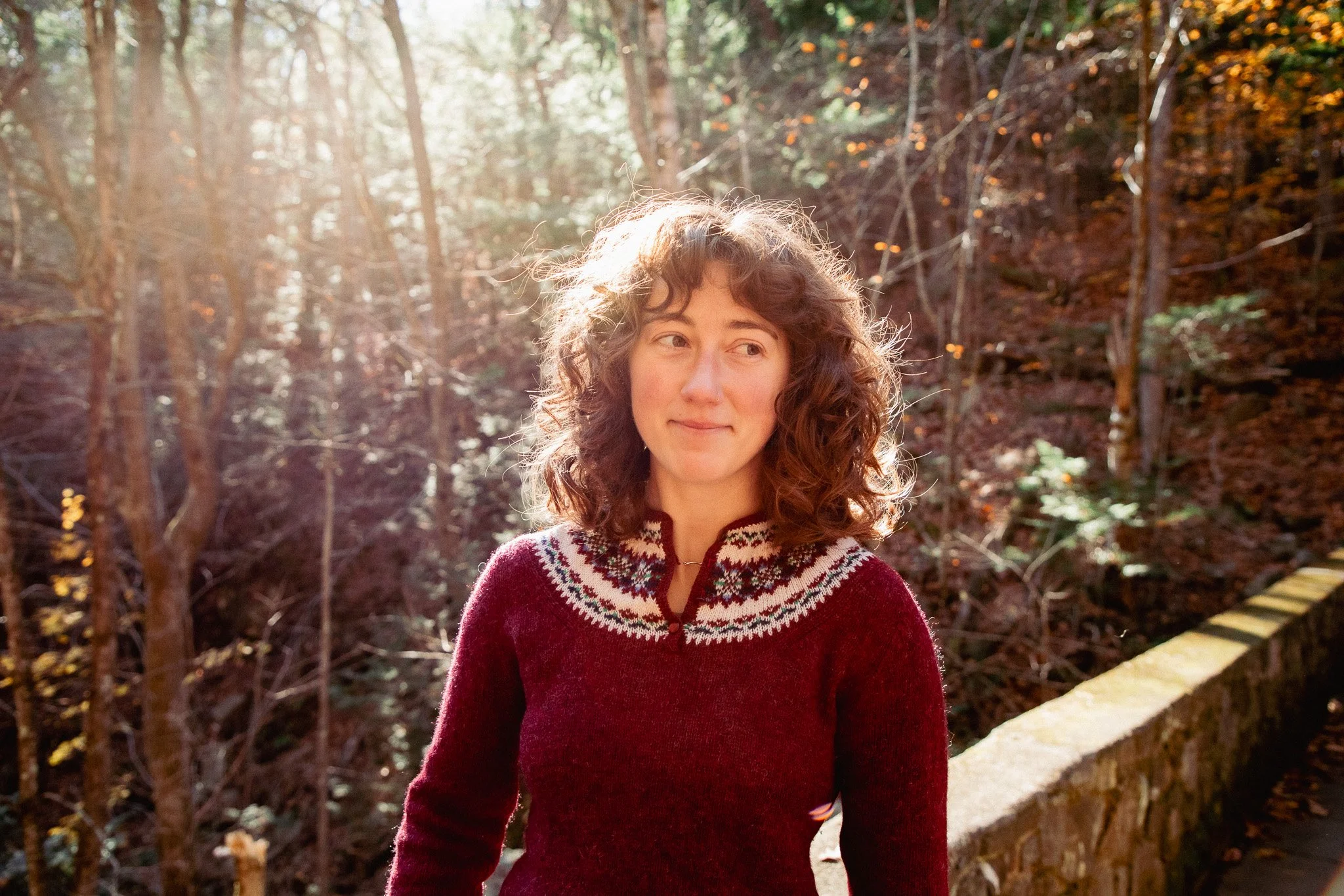 A woman with curly hair wearing a red sweater with white and black patterns standing outdoors in a wooded area with sunlight filtering through trees.
