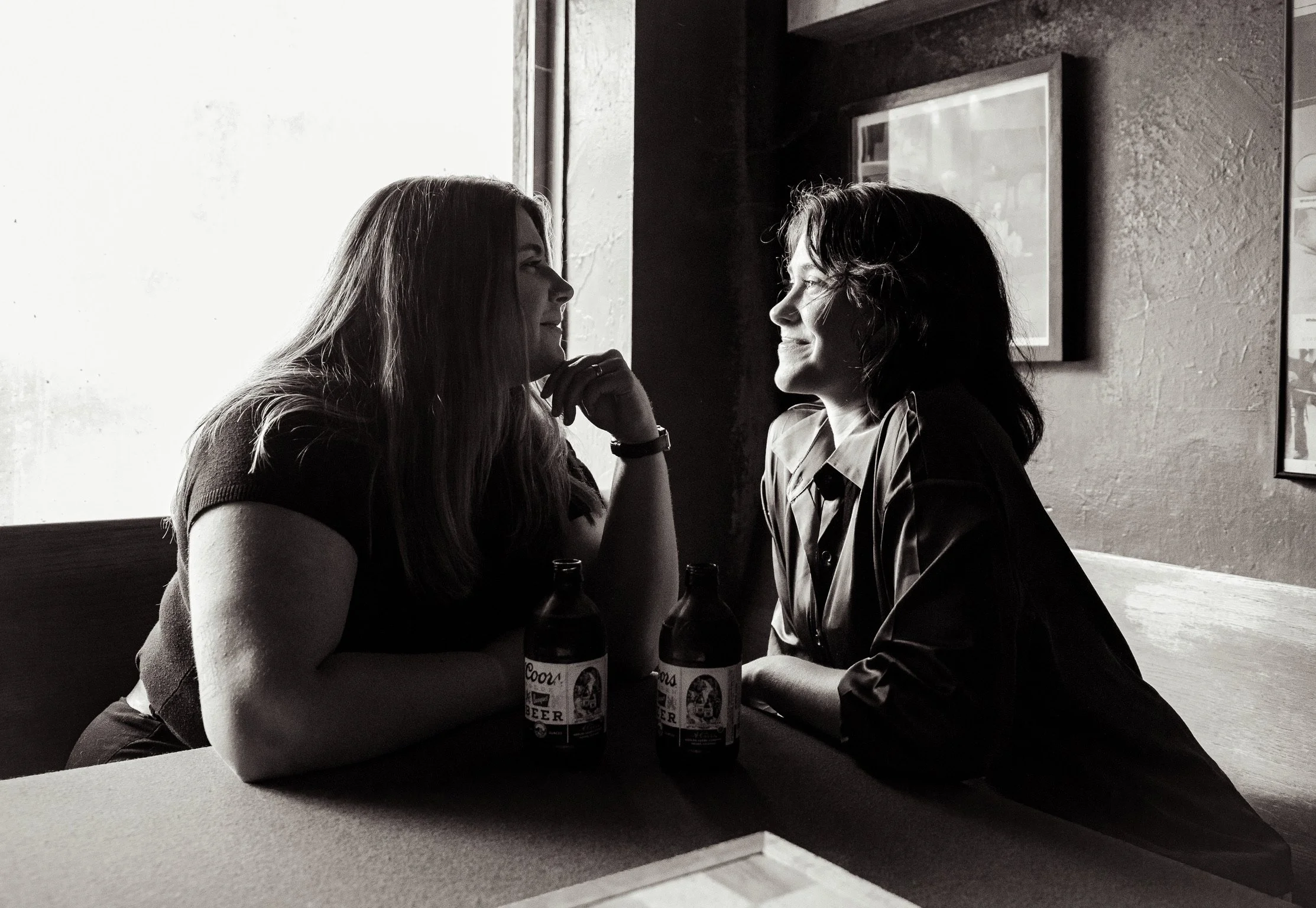Two women are sitting in a bar, facing each other and smiling. They each have a bottle of Coors beer on the table between them. The woman on the left has long hair and is resting her chin on her hand, while the woman on the right has wavy hair and is