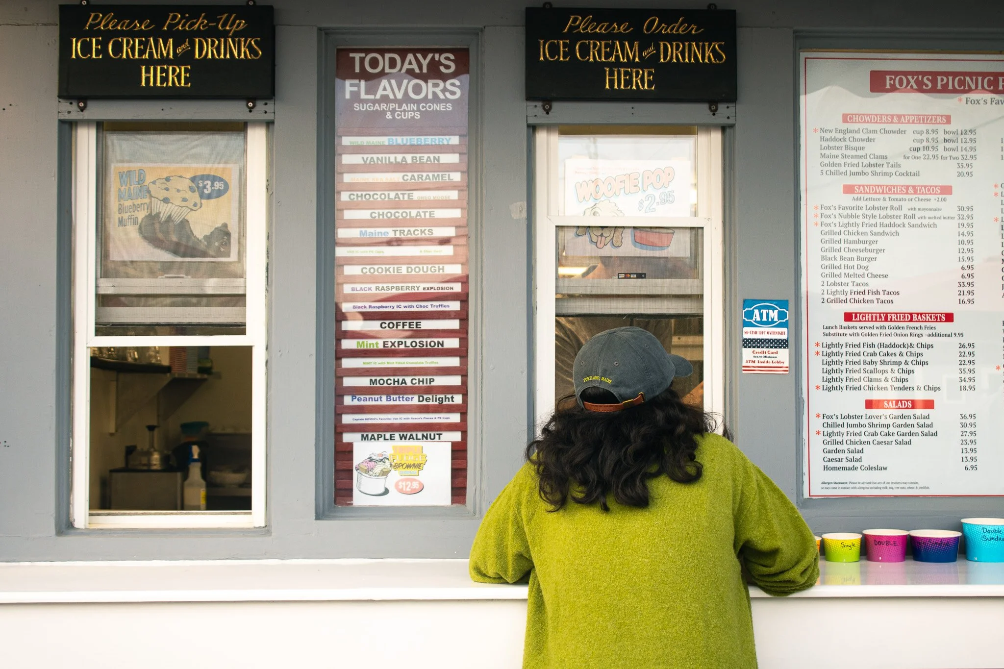 A woman with curly dark hair wearing a bright yellow-green sweater is standing at a counter, facing away from the camera. Behind her, there are signs for ice cream, drinks, and menu items, with a menu board on the right listing various food options l