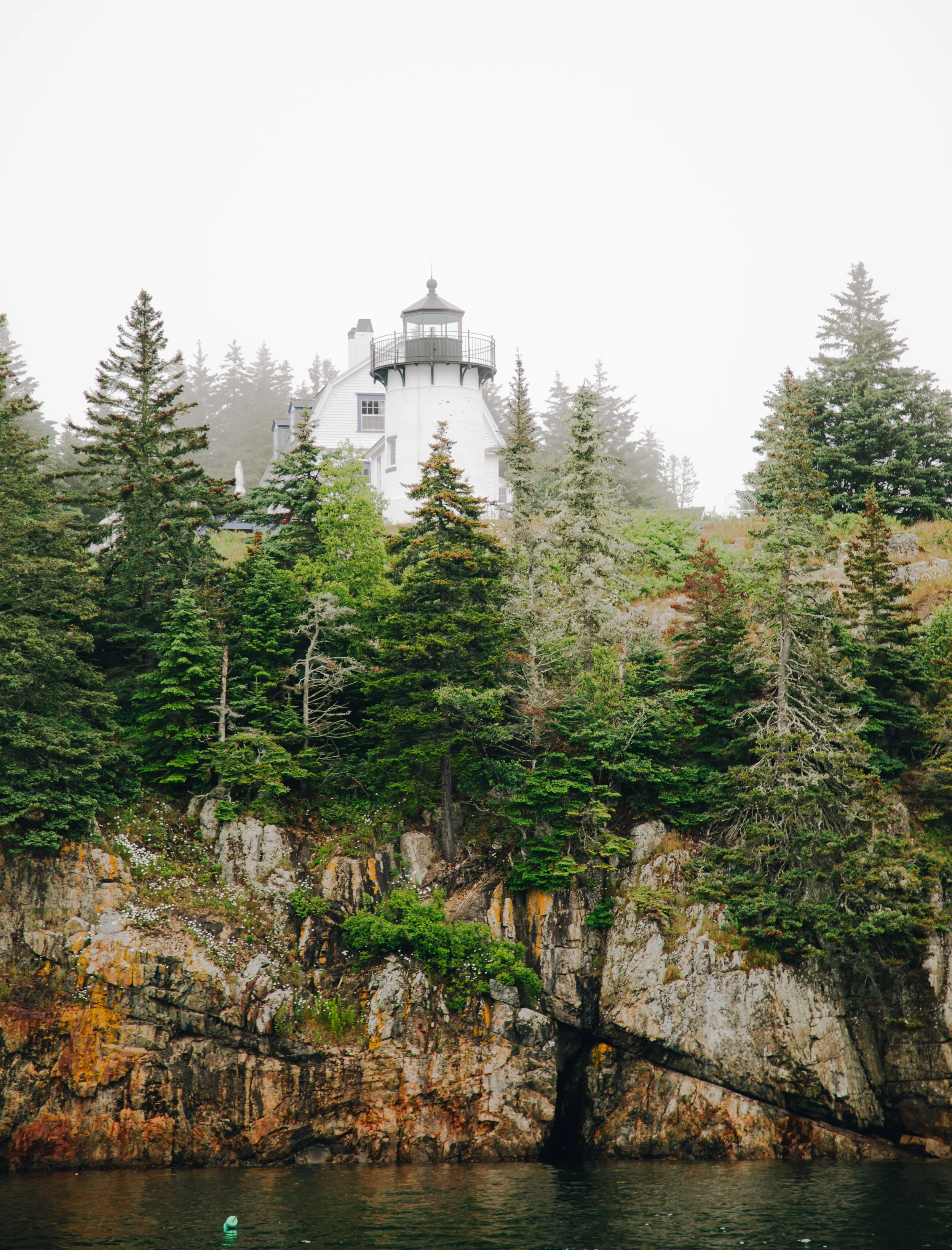 A lighthouse atop a cliff amid tall green trees overlooking a body of water, with a boat floating nearby and foggy weather.