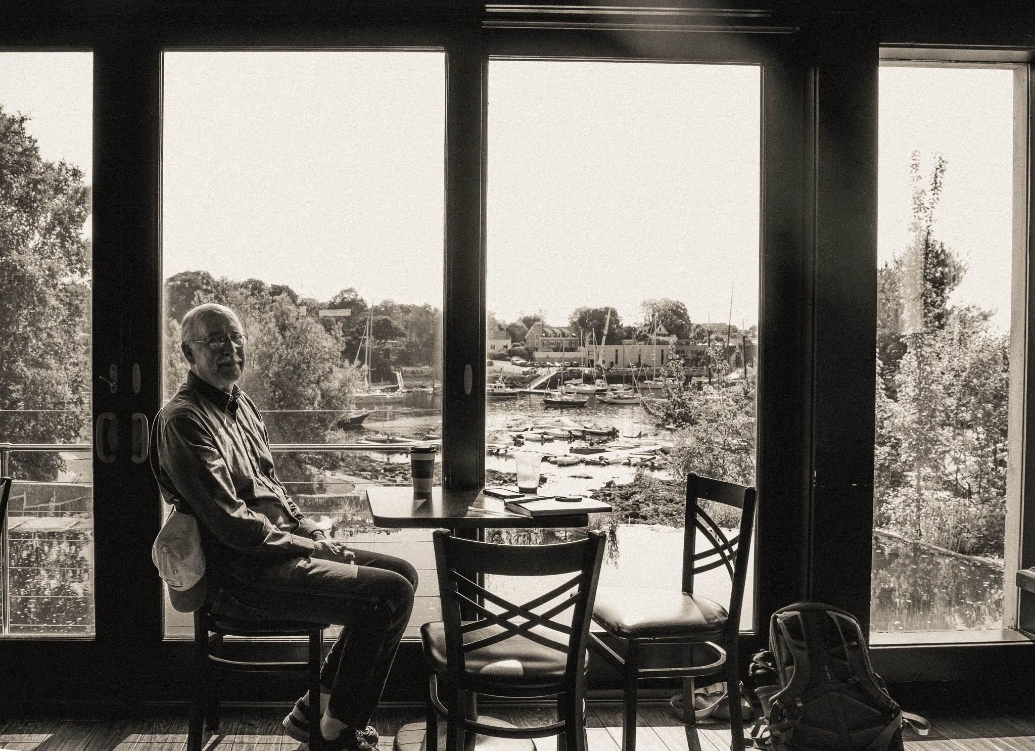 A man sits in a restaurant or cafe near large windows overlooking a marina with boats. The scene is black and white, with natural light coming through the windows, illuminating the man's profile and the interior furniture.