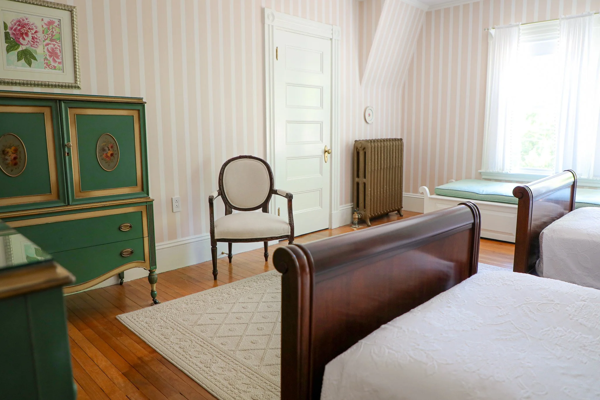 A cozy bedroom with vintage furniture, beige and white striped wallpaper, hardwood floors, a green dresser with floral details, a beige armchair, a radiator near the window, and twin beds with white bedding. Light streams through white curtains.