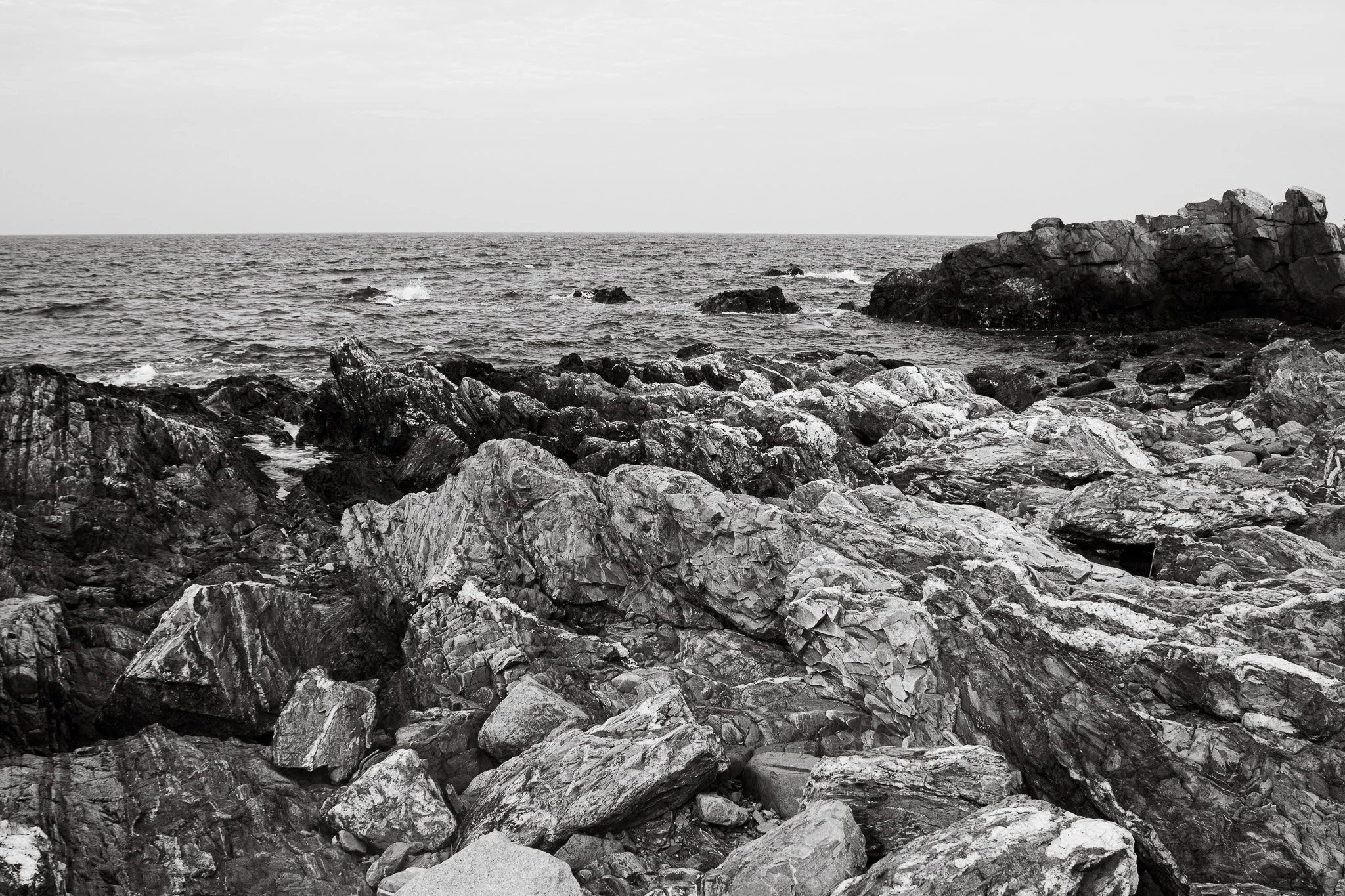 Black and white photograph of a rocky shoreline with waves hitting the rocks and a calm ocean in the background.
