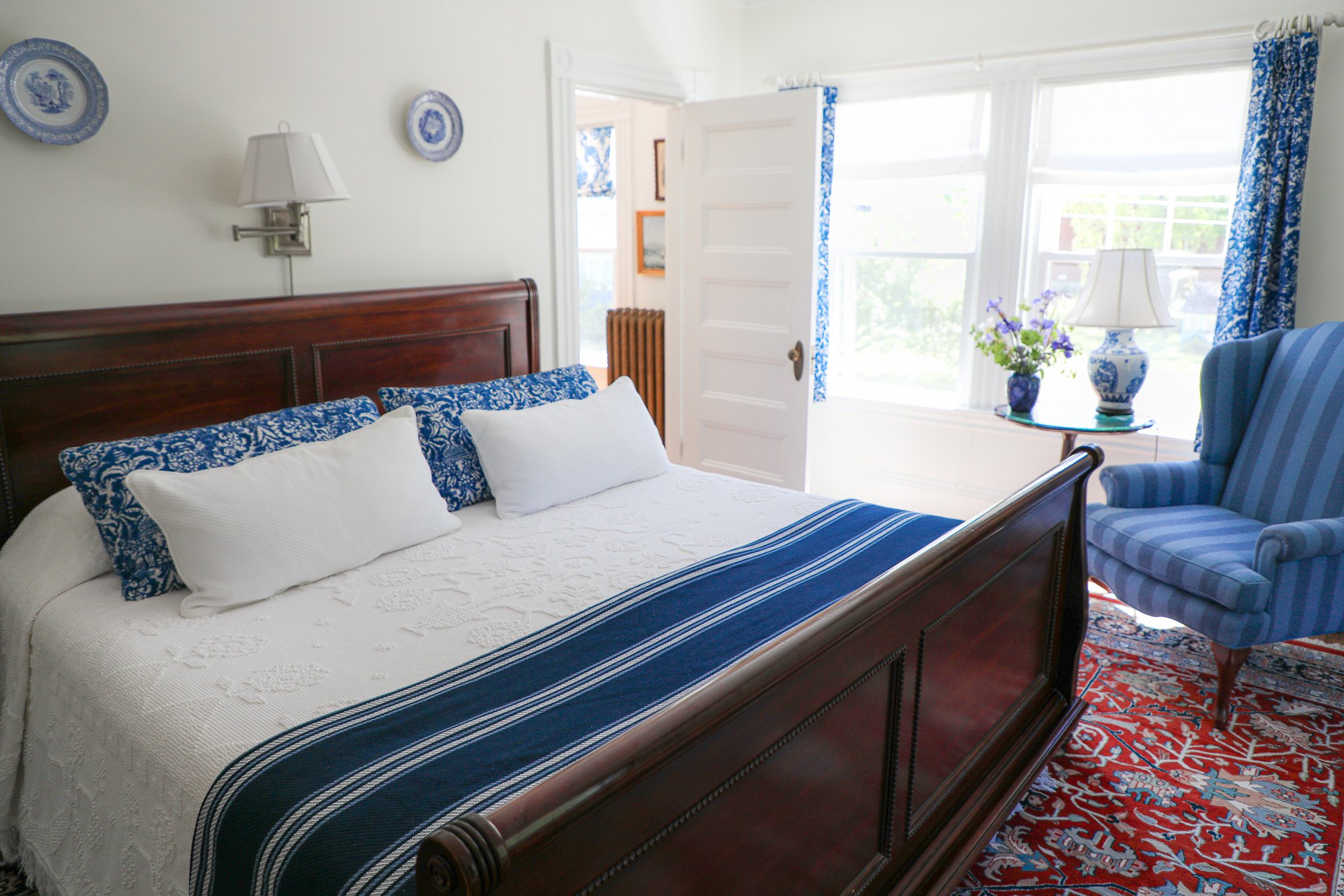 A bedroom with a wooden bed, white bedspread, blue and white pillows, a bedside lamp, decorative blue plates on the wall, a window with blue-patterned curtains, a side table with a vase of purple flowers, and an armchair covered in blue striped fabri