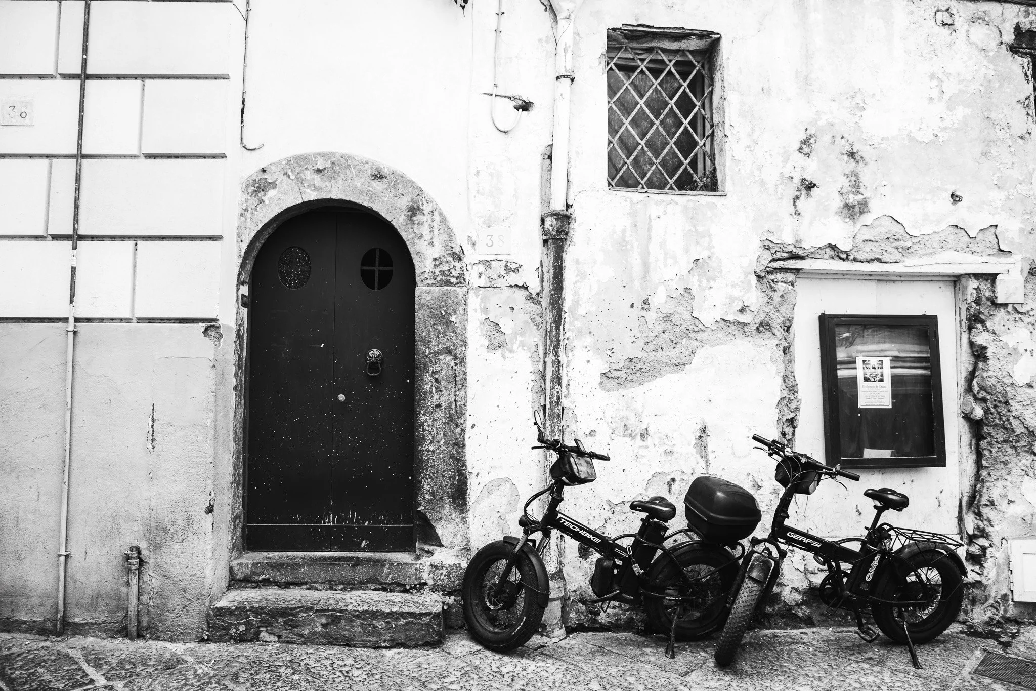 Black bicycles parked outside a weathered, peeling white wall with a black door and a small window with iron bars.