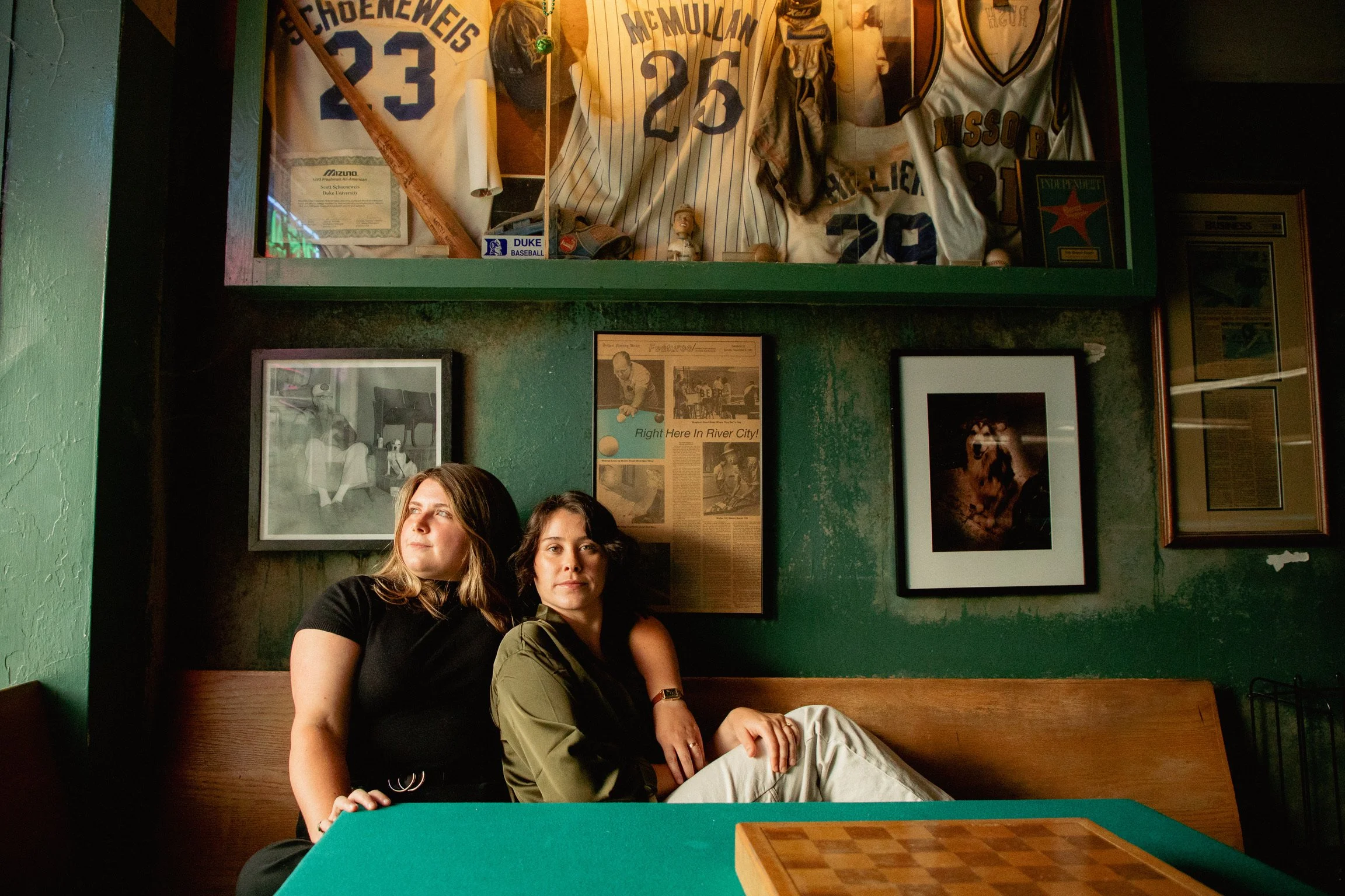 Two women sitting on a wooden bench in a room with green walls and sports memorabilia hanging on the wall behind them. The memorabilia include framed pictures, newspaper clippings, and sports jerseys.