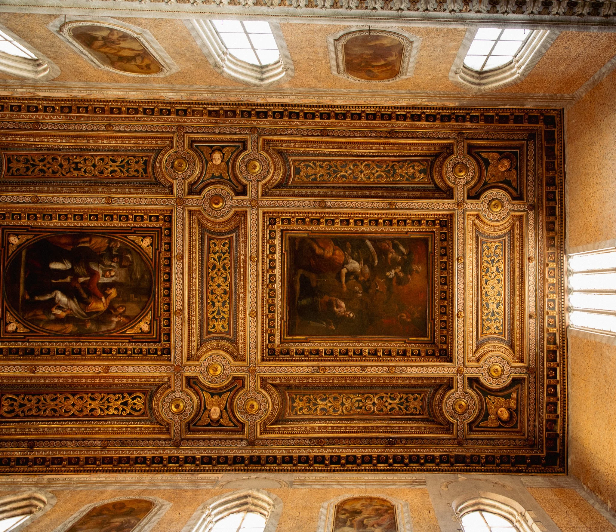 The ceiling of a historic building with ornate gold and painted decorations, including paintings and framed artworks.