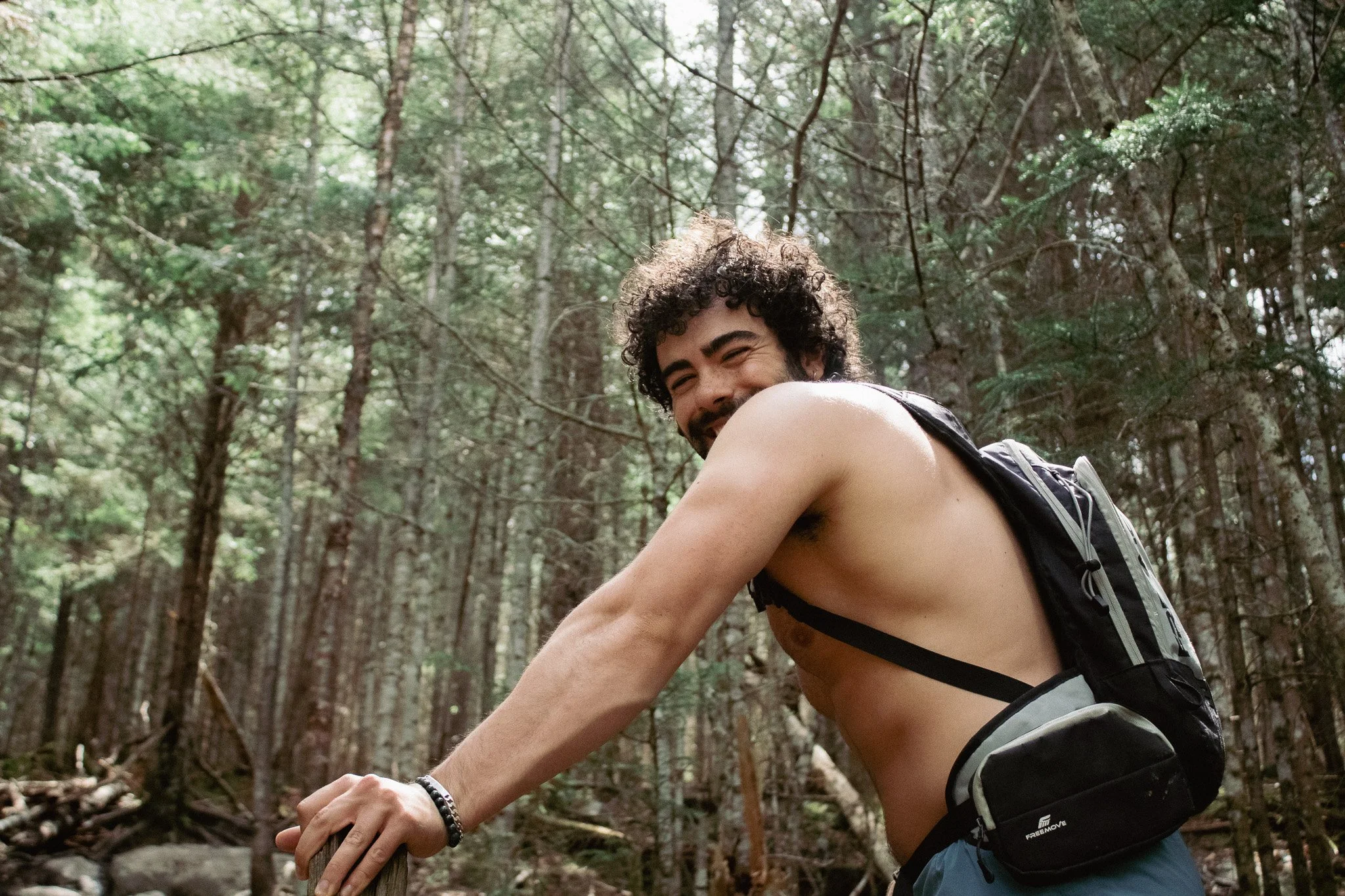 A man with curly hair and a beard smiling while hiking in a forest, wearing a backpack and shorts.
