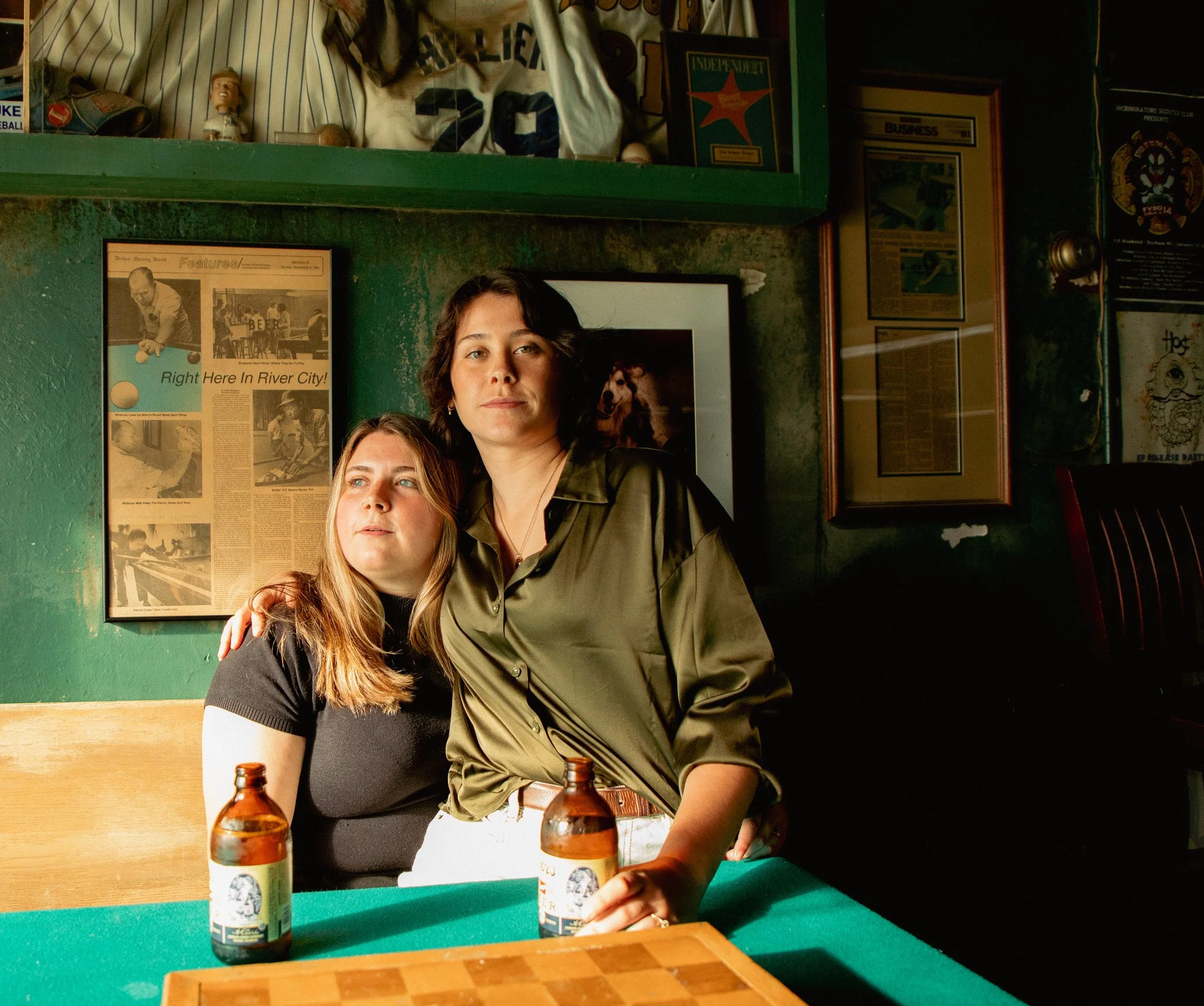 Two women in a bar, one with long blonde hair wearing a black shirt and the other with short dark hair wearing an olive green shirt and crossbody bag, sitting at a table with two beer bottles. The background features vintage posters, framed photos, a