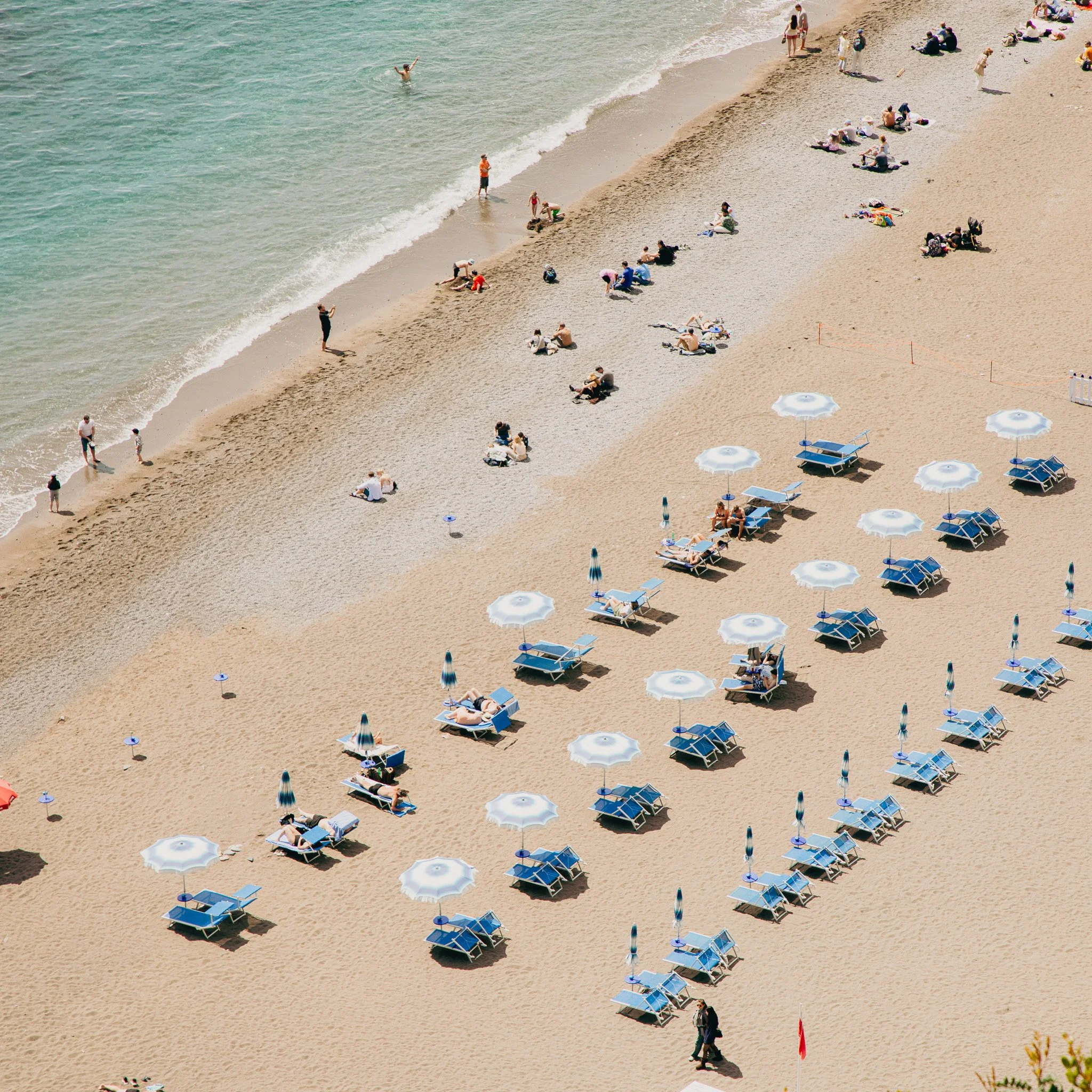 Beach with people sitting on lounge chairs and umbrellas, some walking along the shoreline, and others swimming in the ocean on a sunny day.