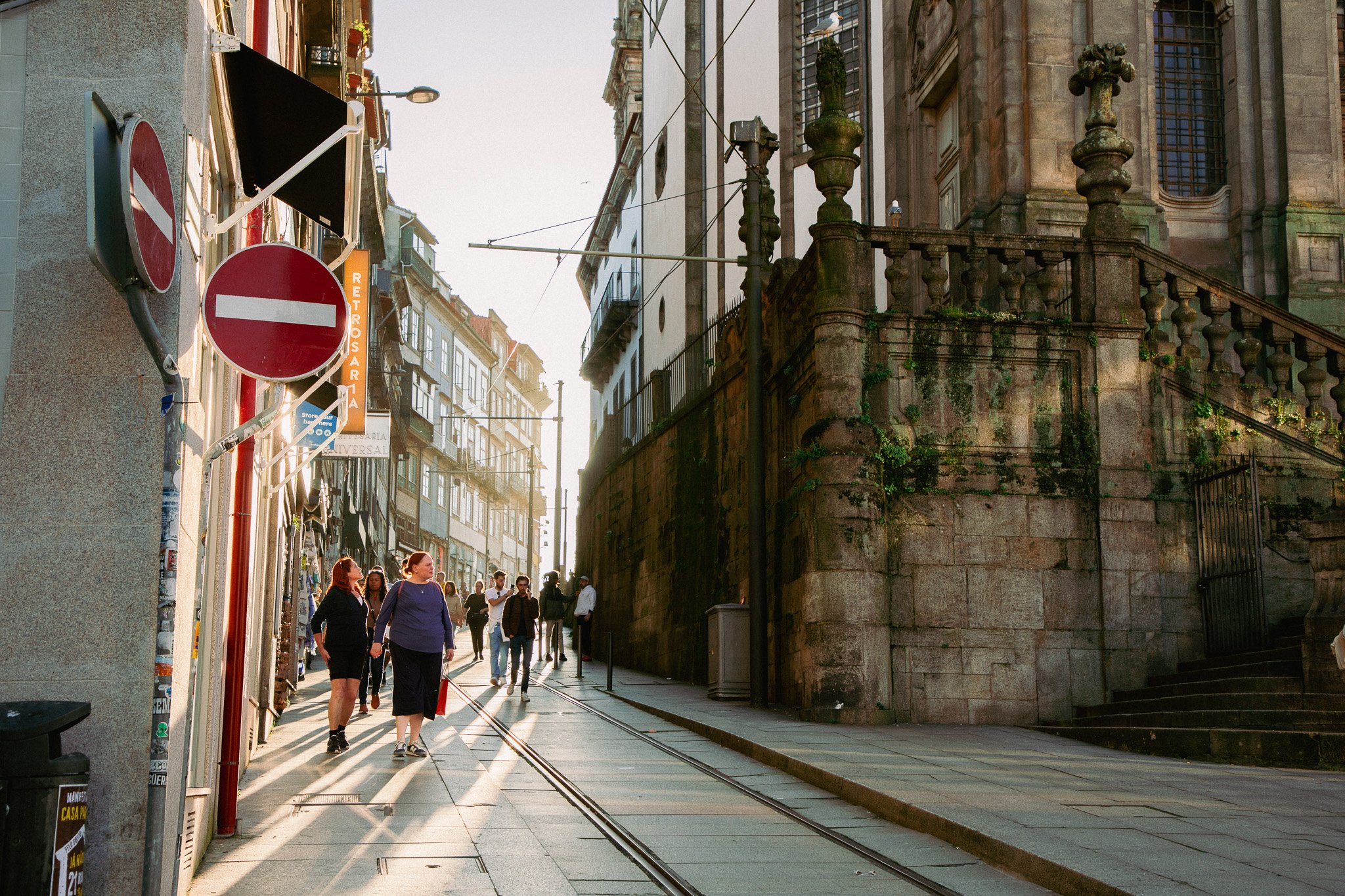 A busy city street scene with pedestrians walking along the sidewalk. There are old stone buildings with balconies and some greenery visible. Tram tracks run along the street, and various signs, including a no-entry sign, are attached to poles. The s