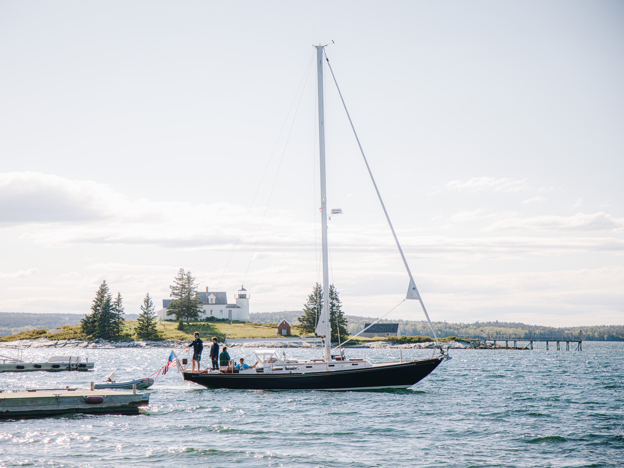 A sailboat with four people on board, docked at a marina with a lighthouse and trees in the background, under a cloudy sky.